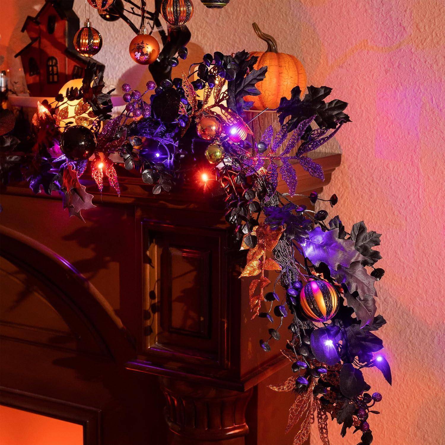 Eine dekorierte Halloween-Laternenschau mit leuchtenden violett-orangefarbenen Lichtern, schwarzen Blättern und hängenden Ornamenten auf einem Kaminsims vor einer rosa Wand.