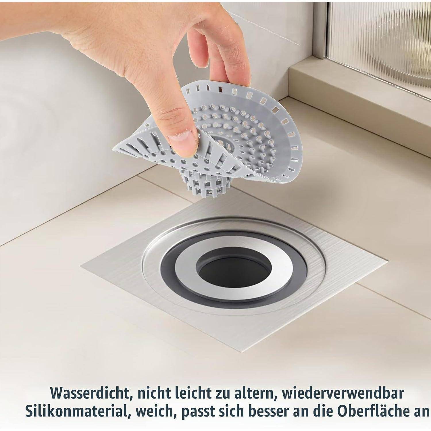 Hand hebt eine graue Silikon-Ablaufabdeckung über einen Bodenablauf in einem Dusch- oder Spülbereich; deutscher Text besagt, dass sie wasserdicht, langlebig, wiederverwendbar und weich ist.