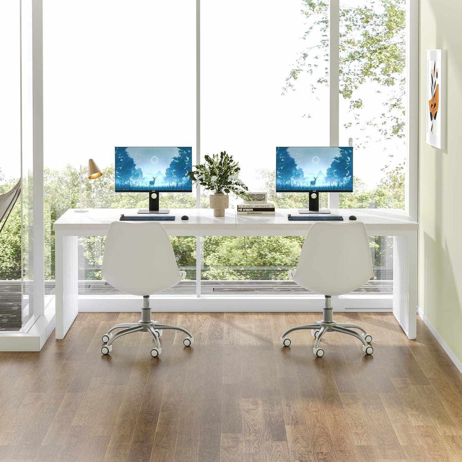Helles Homeoffice mit zwei weißen Schreibtischen, passenden Stühlen, zwei Computermonitoren und einer zentralen Pflanze neben großen Fenstern mit Ausblick auf Bäume.