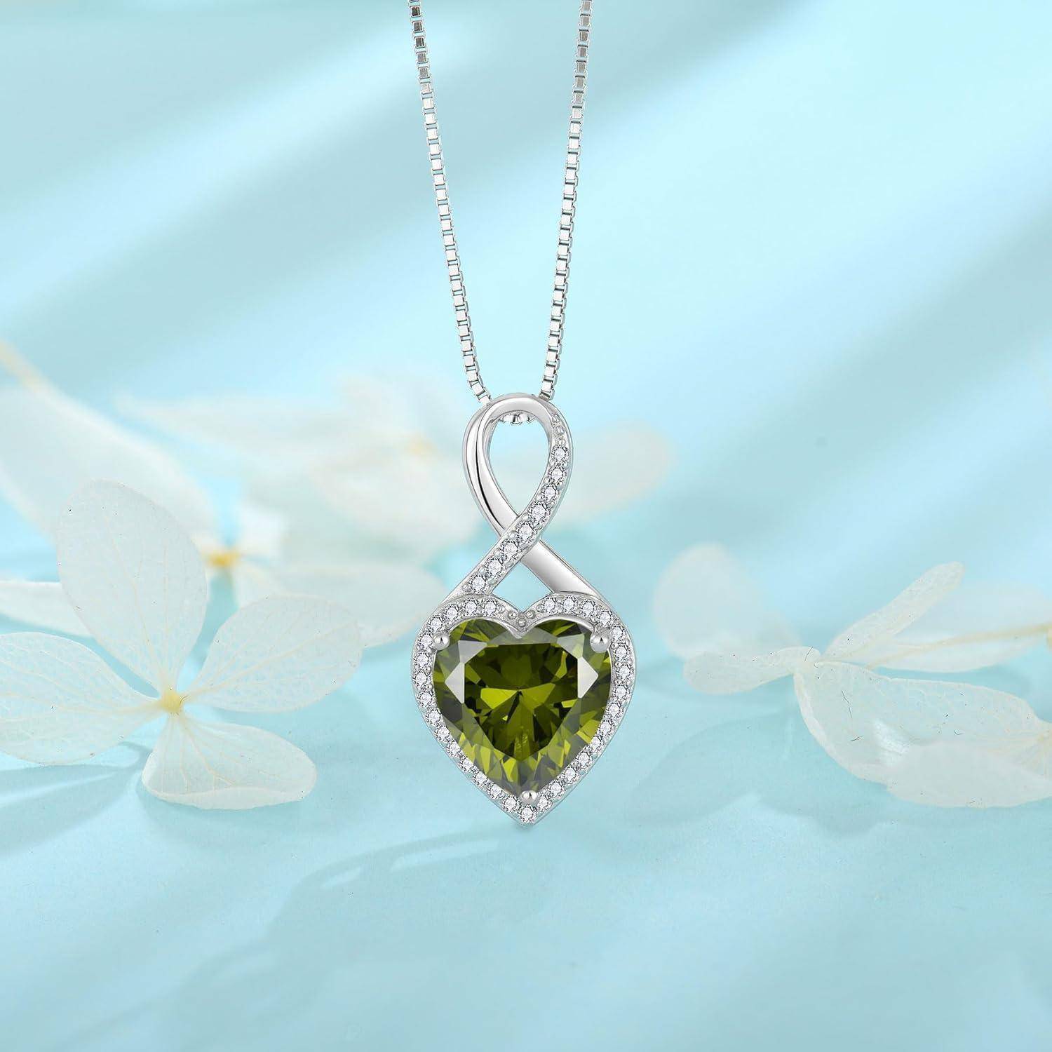 Silbernes Herz-Anhänger-Collier mit einer Unendlichkeitsschleife und grünem herzförmigem Edelstein, akzentuiert durch kleine klare Steine auf einem sanft blauen floralen Hintergrund.