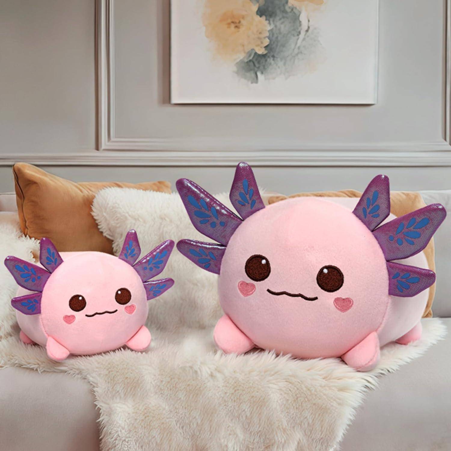 Zwei Plüsch-Axolotl-Spielzeuge, eines groß und eines klein, sitzen auf einem Sofa mit Kissen und einer Decke in einem gemütlichen Wohnzimmer.
