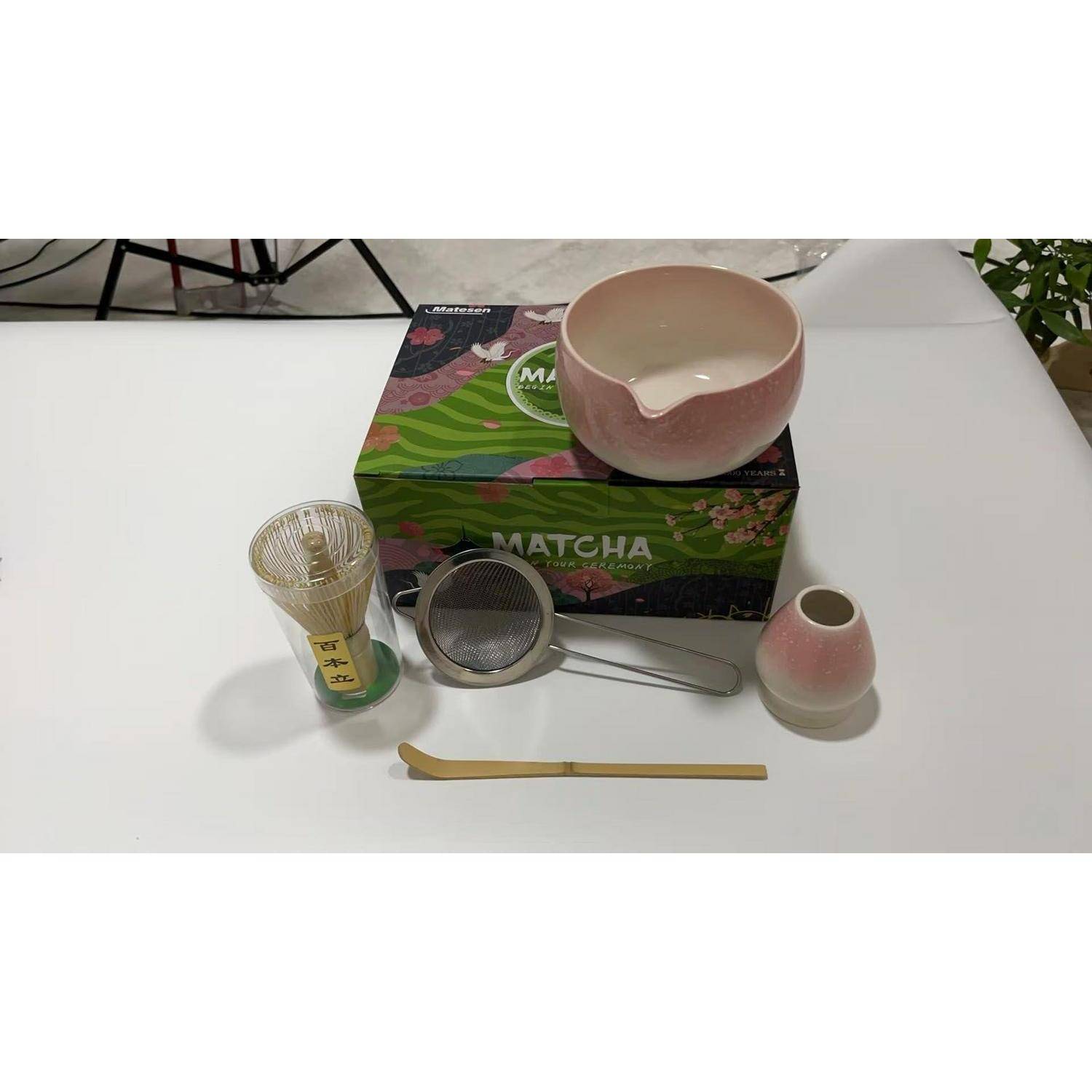 Matcha-Tee-Set auf einem Tisch: rosa Schale mit Ausguss, Schneebesen, Sieb, Messlöffel und Verpackungsbox.