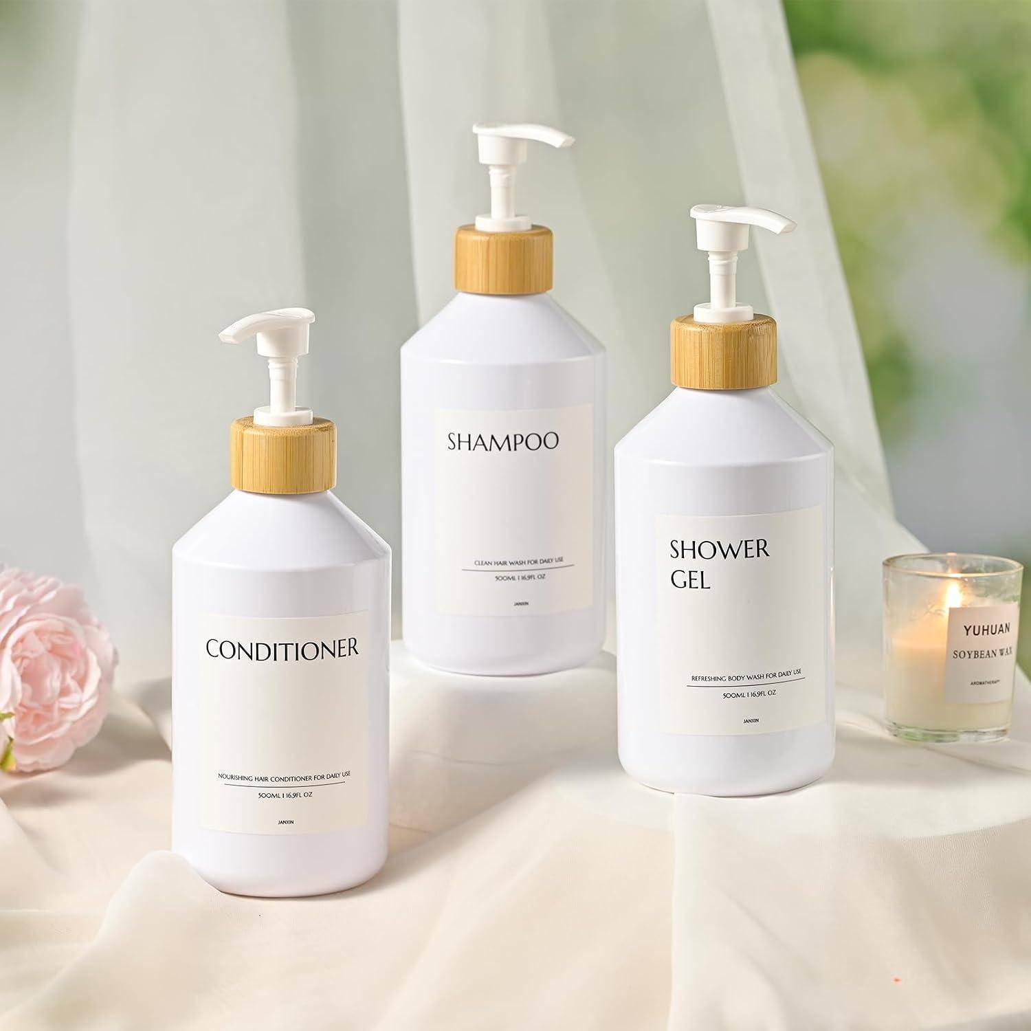 Drei weiße Pumpflaschen mit den Aufschriften „Conditioner", „Shampoo"" und „Duschgel"" auf einem weichen Stoff-Hintergrund neben einer Kerze und einer Blume.