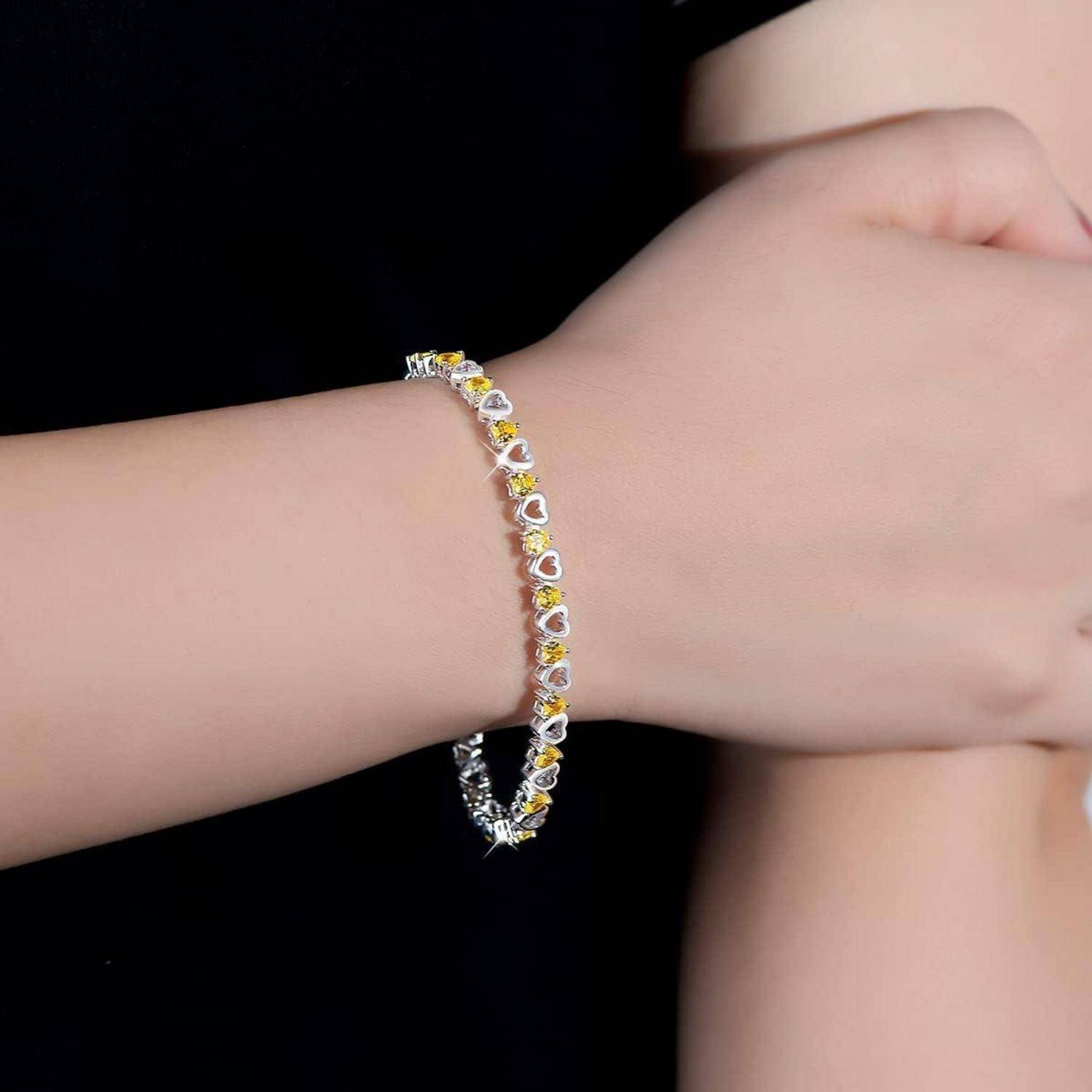 Nahaufnahme eines Handgelenks, das ein zartes silbernes Armband mit herzförmigen gelb-transparenten Steinen vor schwarzem Hintergrund trägt.