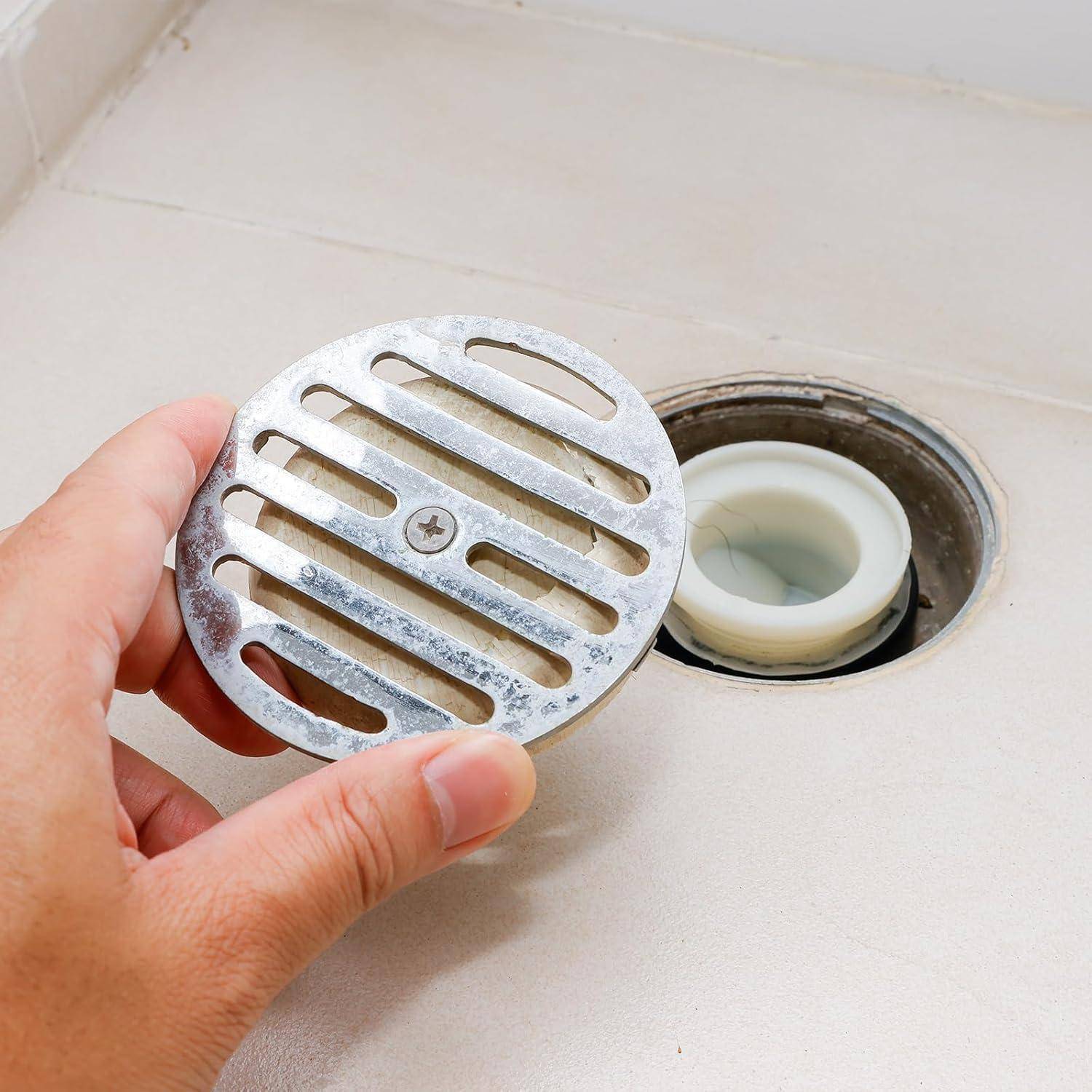 Hand, die ein Metallkanaldeckel über einem offenen Bodenablauf mit einer weißen Rohreinsatz hält.