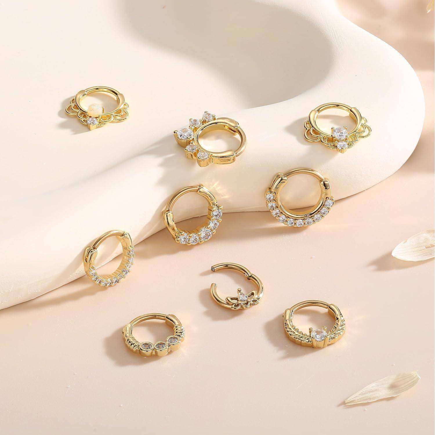 Acht goldene Creolen mit funkelnden klaren Steinen sind auf einer hellcremefarbenen Oberfläche angeordnet und zeigen verschiedene dekorative Designs.
