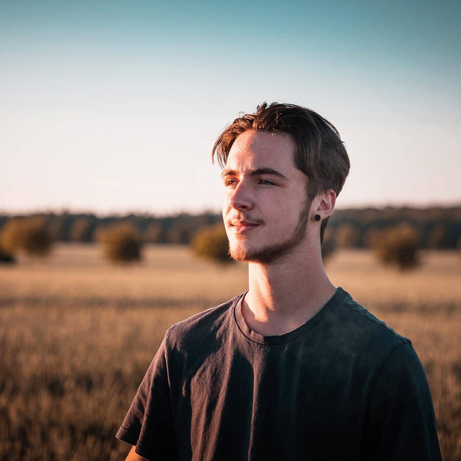 Ein junger Mann in einem schwarzen T-Shirt steht in einem sonnenbeschienenen Feld und blickt bei Sonnenuntergang zur Seite.