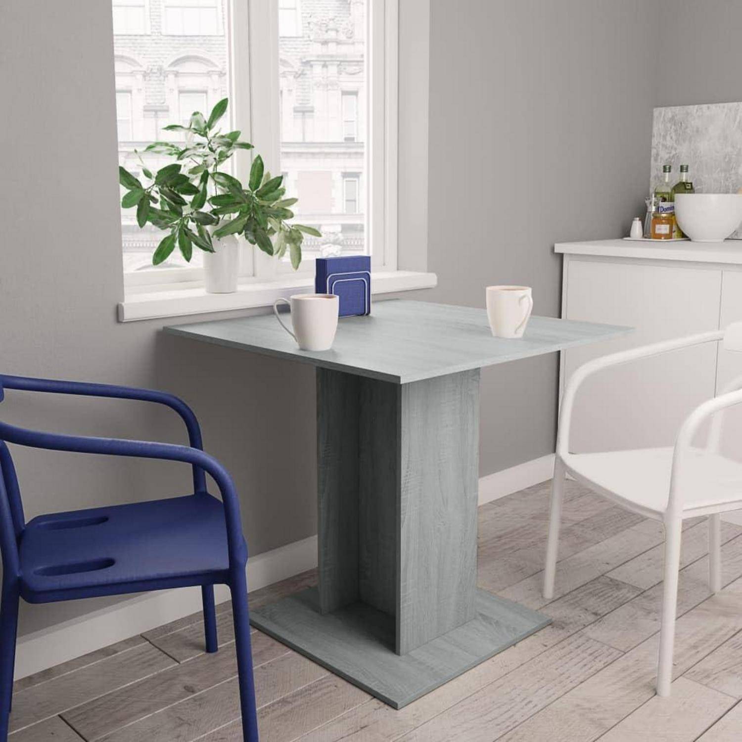 Kleiner moderner Essbereich mit einem grauen Sockel-Esstisch, blau-weißen Stühlen, zwei Tassen, einer Pflanze am Fenster und einem weißen Sideboard.