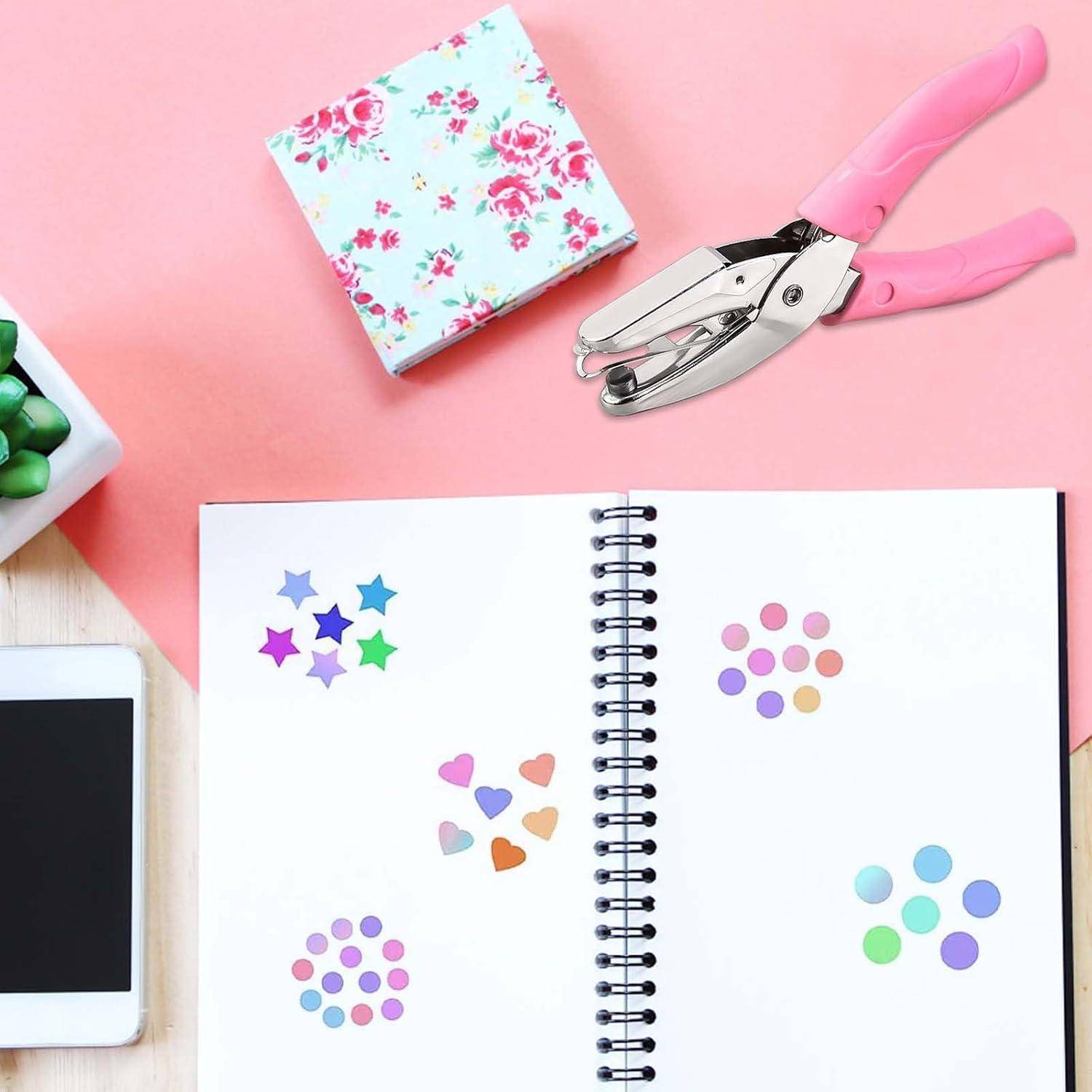 Draufsicht auf ein offenes Spiralnotizbuch mit bunten Aufklebern, neben einem rosa Bastellocher und einer Blumenschachtel auf einem rosa Schreibtisch.