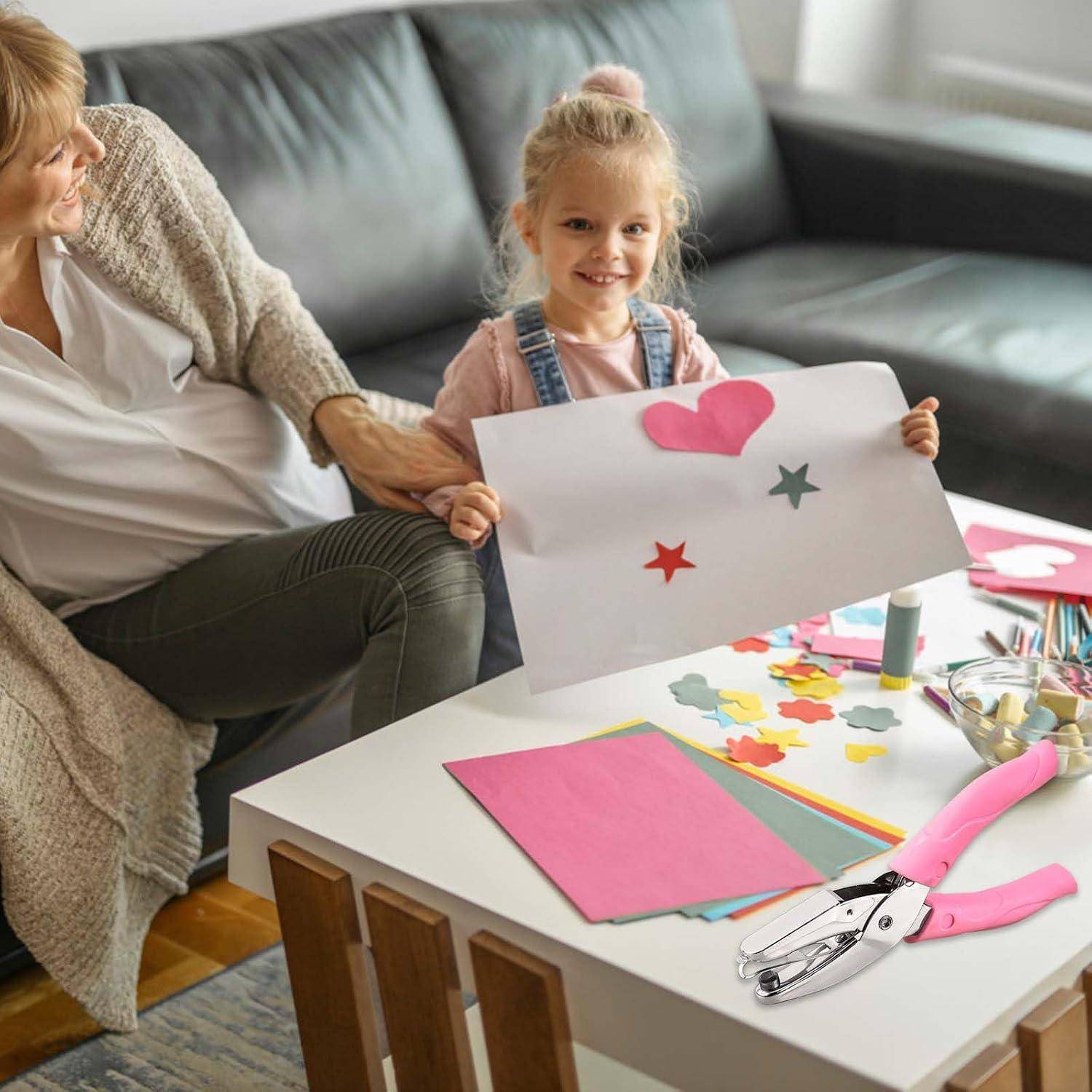 Junges Mädchen zeigt stolz ein verziertes Papierhandwerk, während es mit einer älteren Frau an einem Basteltisch mit farbenfrohen Ausschnitten und Werkzeugen sitzt.