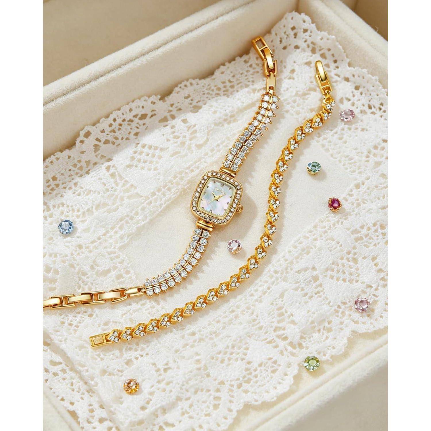 Goldarmbanduhren und passender Kristallarmband auf weißer Spitze in einer cremefarbenen Schmuckschatulle, mit verstreuten farbigen Edelsteinen.