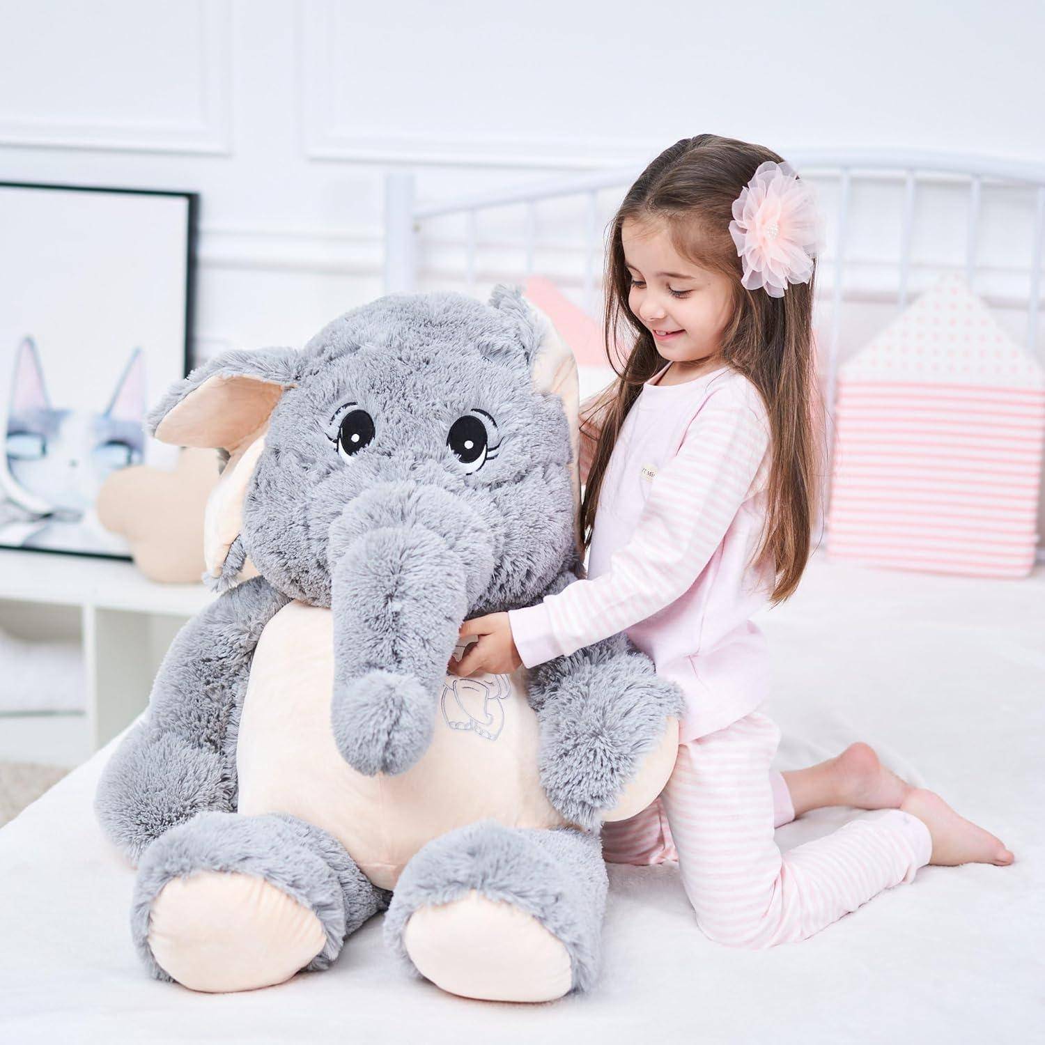 Junges Mädchen in rosa Pyjama kniet auf einem Bett und umarmt ein großes graues Plüschelefantenspielzeug.