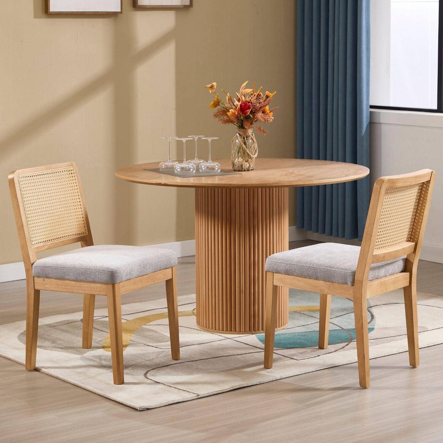Runder Holzesstisch mit zwei gepolsterten Stühlen mit Rohrgeflecht-Rückenlehne in einem hellen modernen Raum, mit einer Vase mit Blumen und Gläsern auf dem Tisch.
