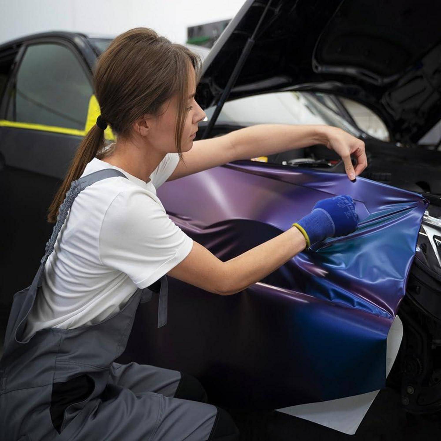 Techniker, der eine schillernde Vinyl-Folie auf einer Motorhaube eines Autos in einer Werkstatt anbringt und die Folie von Hand glättet.