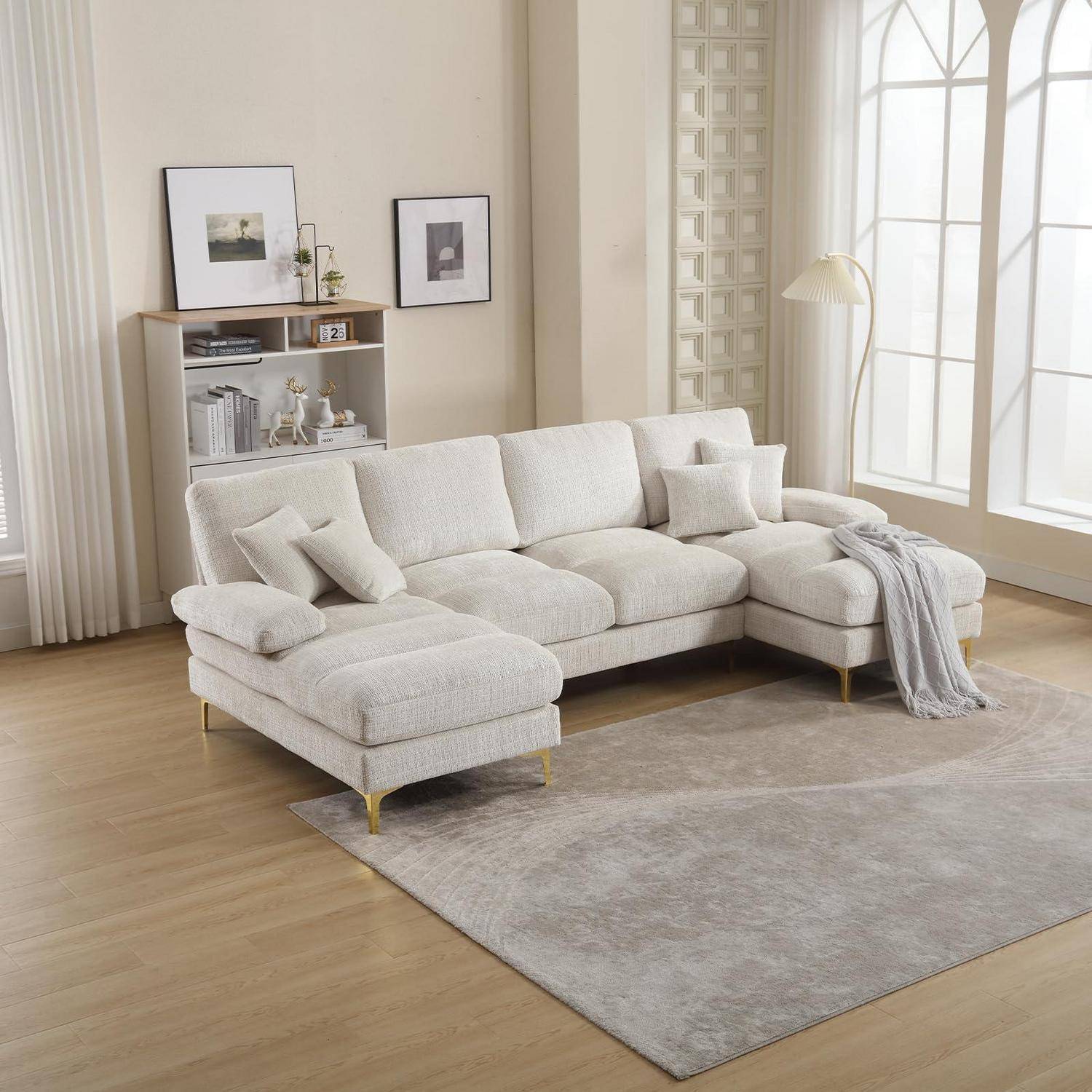 Helles beige L-förmiges Sectional-Sofa mit Kissen und einer Wurfdecke in einem hellen, minimalistischen Wohnzimmer mit Teppich, Regal und hohem Fenster.