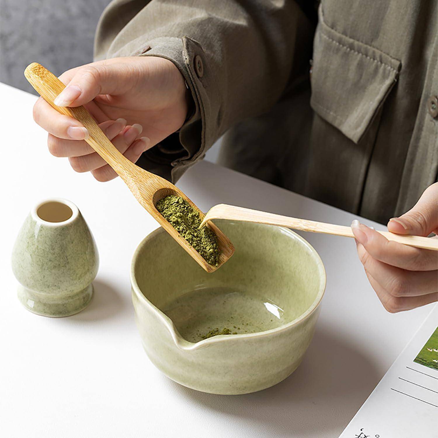 Hände bereiten Matcha-Tee vor, übertragen grünes Teepulver mit einem Holzlöffel in eine Keramikschale mit einem Bambusquirl in der Nähe.