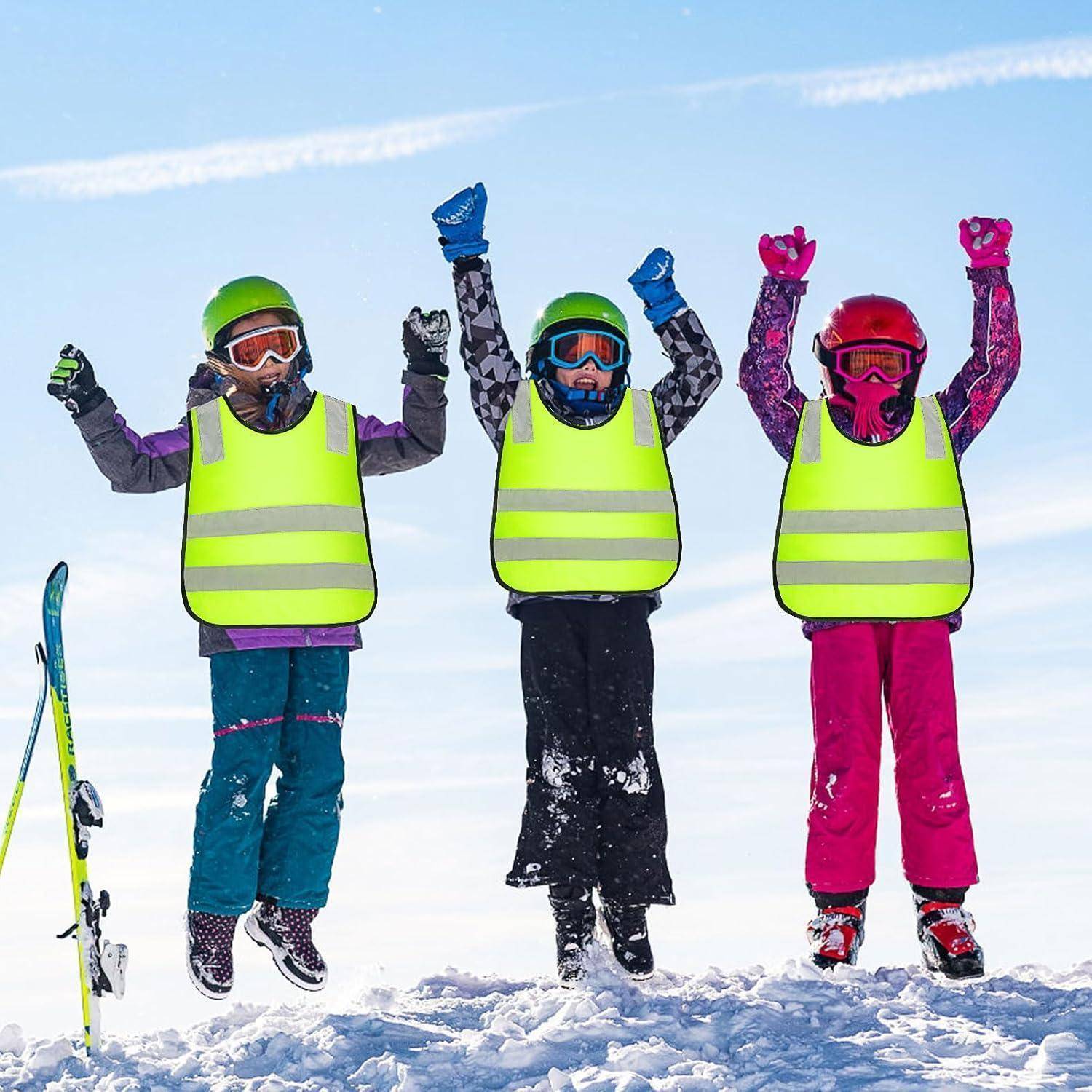 Drei Kinder in Skiausrüstung springen mit erhobenen Armen im Schnee, tragen leuchtend farbige Sicherheitswesten, Helme und Skibrillen neben einem Ski an einem schneebedeckten Hang.