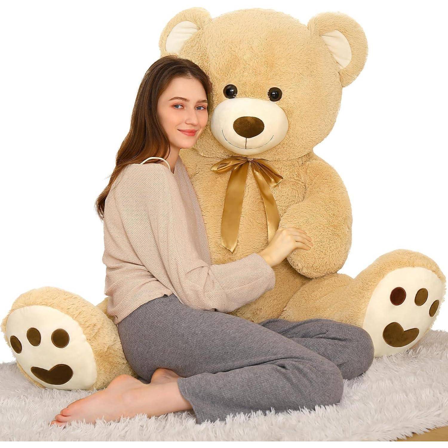 Junge Frau, die sitzt und einen riesigen beige Teddybären mit einer Schleife auf einem weißen Teppich umarmt.