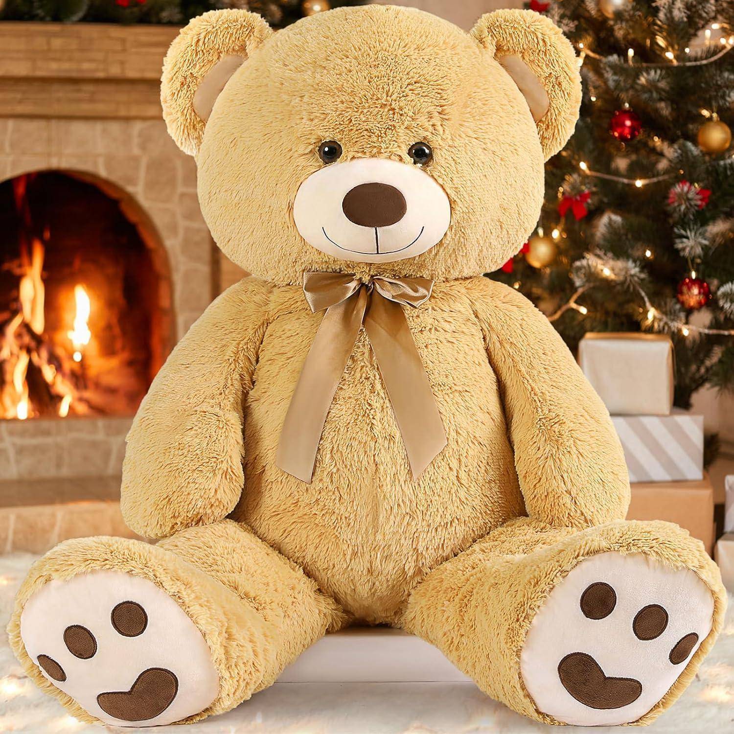 Ein großer beiger Teddybär mit einer Schleife sitzt vor einem brennenden Kamin und einem geschmückten Weihnachtsbaum.