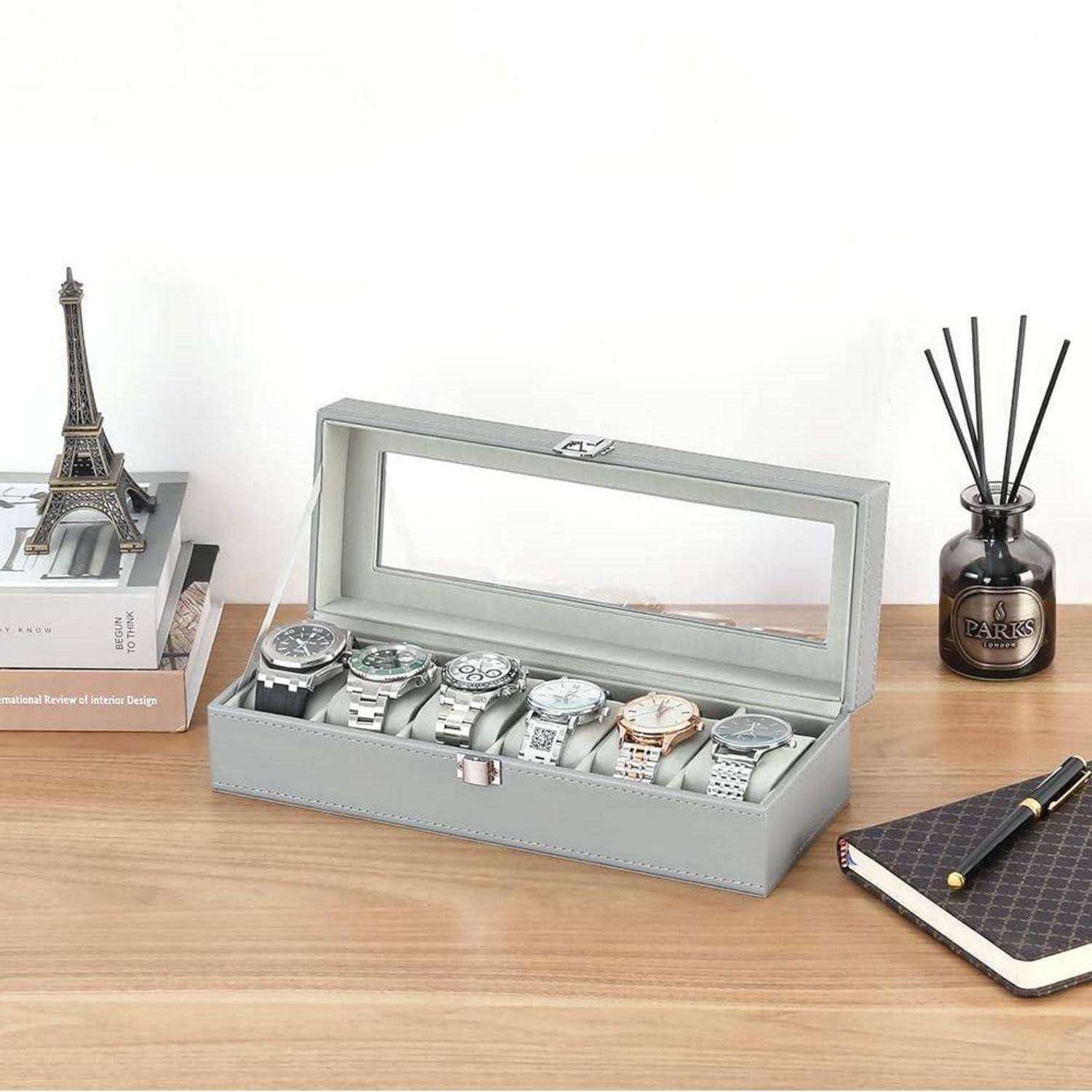 Sechs Uhren, die in einer Vitrine mit grauer Glasplatte auf einem Holzschreibtisch ausgestellt sind, mit einem kleinen Eiffelturm-Modell, Büchern, einem Raumduft-Diffusor und einem Notizbuch in der Nähe.
