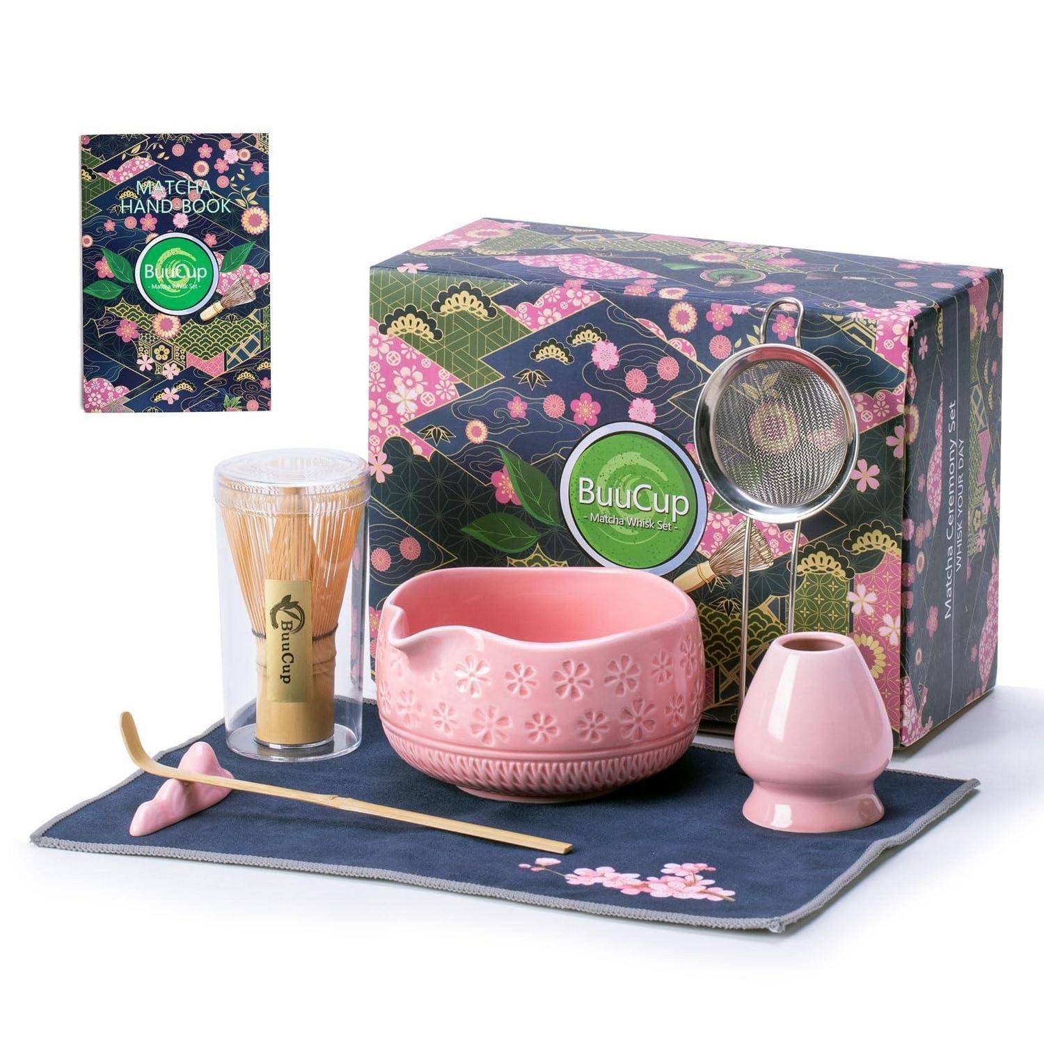 Matcha-Tee-Set Geschenkbox mit Handbuch, Schneebesen in durchsichtigem Becher, rosa Schale und Löffel, Sieb und kleine Vase auf einer Stoffmatte.