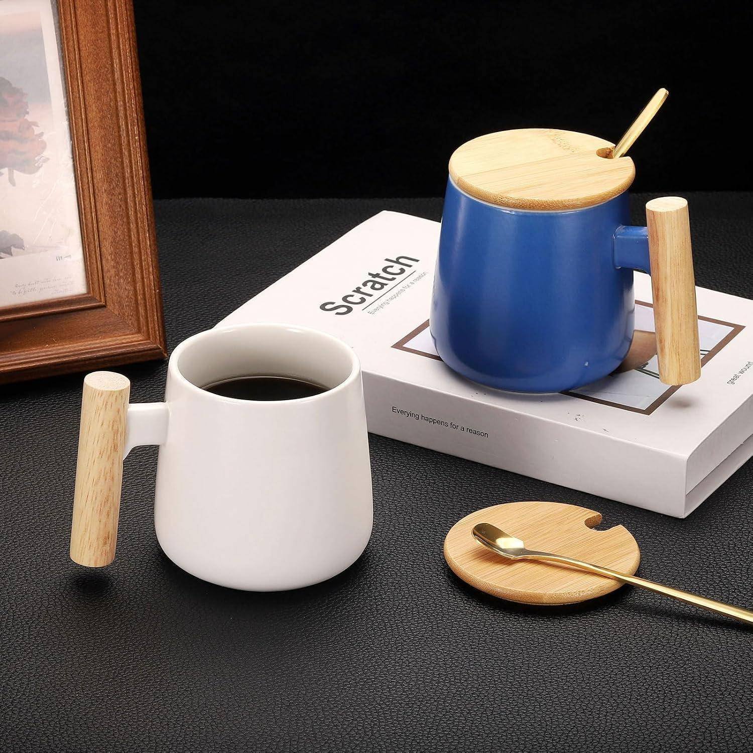 Zwei Keramiktassen mit Holzgriffen und Bambusdeckeln, eine weiße mit Kaffee und eine blaue auf einem Buch, neben einem Löffel auf einem Deckeluntersetzer.
