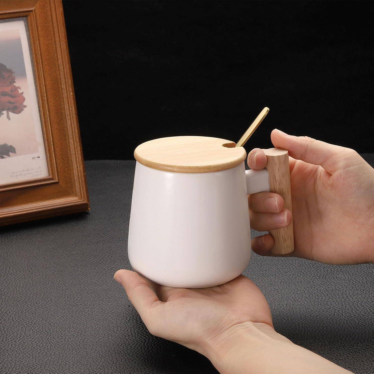 Eine Hand hält eine weiße Keramiktasse mit einem Holzdeckel und Löffel, während eine andere Hand sie gegen einen dunklen Tischplatzhintergrund abstützt.