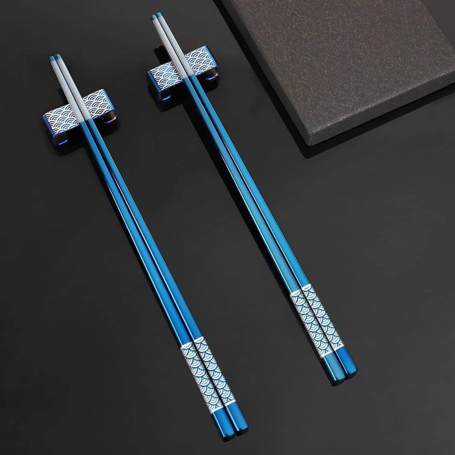 Zwei Paar blaue Essstäbchen mit gemusterten Silberenden, auf einer dunklen Oberfläche neben einem schwarzen Tablett angeordnet.