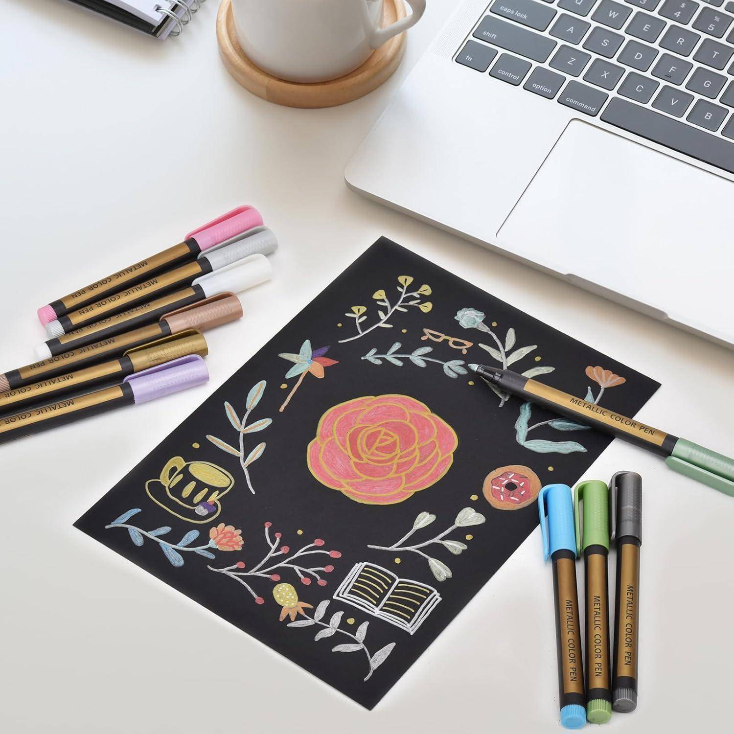 Eine schwarze Karte zeigt ein handgezeichnetes Blumenmotiv mit einer zentralen rosa Rose, umgeben von Blättern, Schmetterlingen und kleinen Symbolen, mit Farbstiften und einem Laptop in der Nähe.