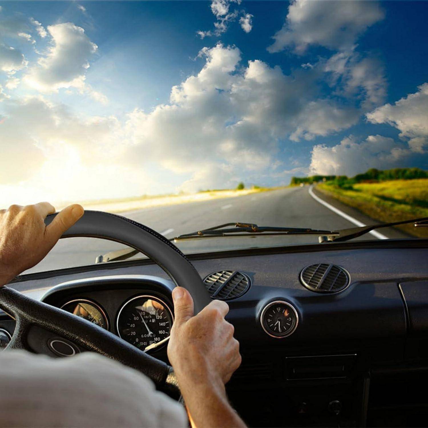 Ansicht des Fahrers aus dem Inneren eines Autos, das auf einer ländlichen Straße unter einem hellen, bewölkten Himmel fährt, mit Händen am Lenkrad.