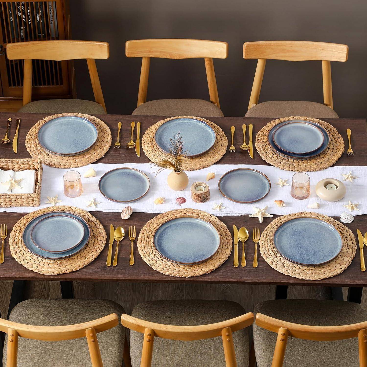 Elegant gedeckter Esstisch für eine Mahlzeit, mit blauen Tellern auf geflochtenen Untersetzer, goldenem Besteck und Stühlen um einen neutralen Tischläufer herum mit Kerzen und Dekorationen.