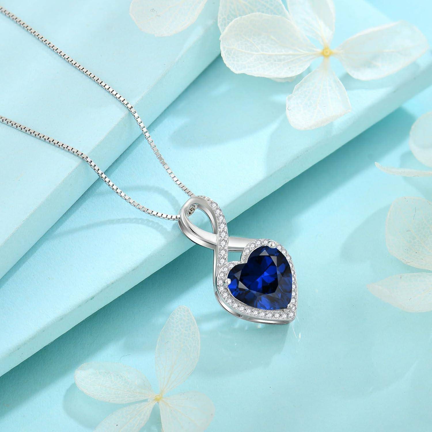Silberkette mit herzförmigem blauem Edelstein-Anhänger, umrahmt von kleinen klaren Steinen, auf hellblauem Blumenhintergrund.