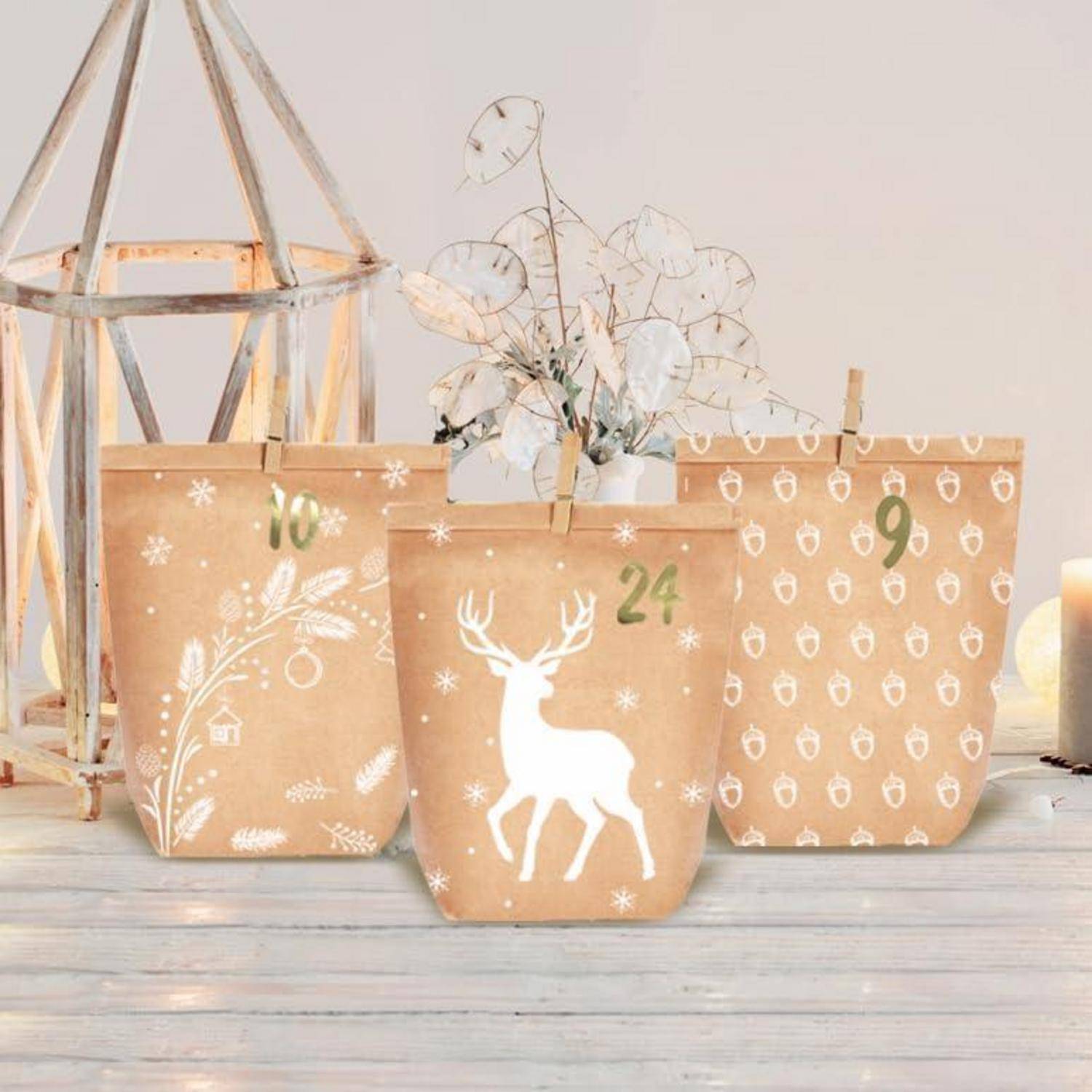 Drei Kraftpapier-Adventskalender-ähnliche Geschenktüten mit weißen Weihnachtsdesigns und Wäscheklammern, eine mit der Nummer 24 gekennzeichnet, auf einer hellen Holzoberfläche angeordnet.