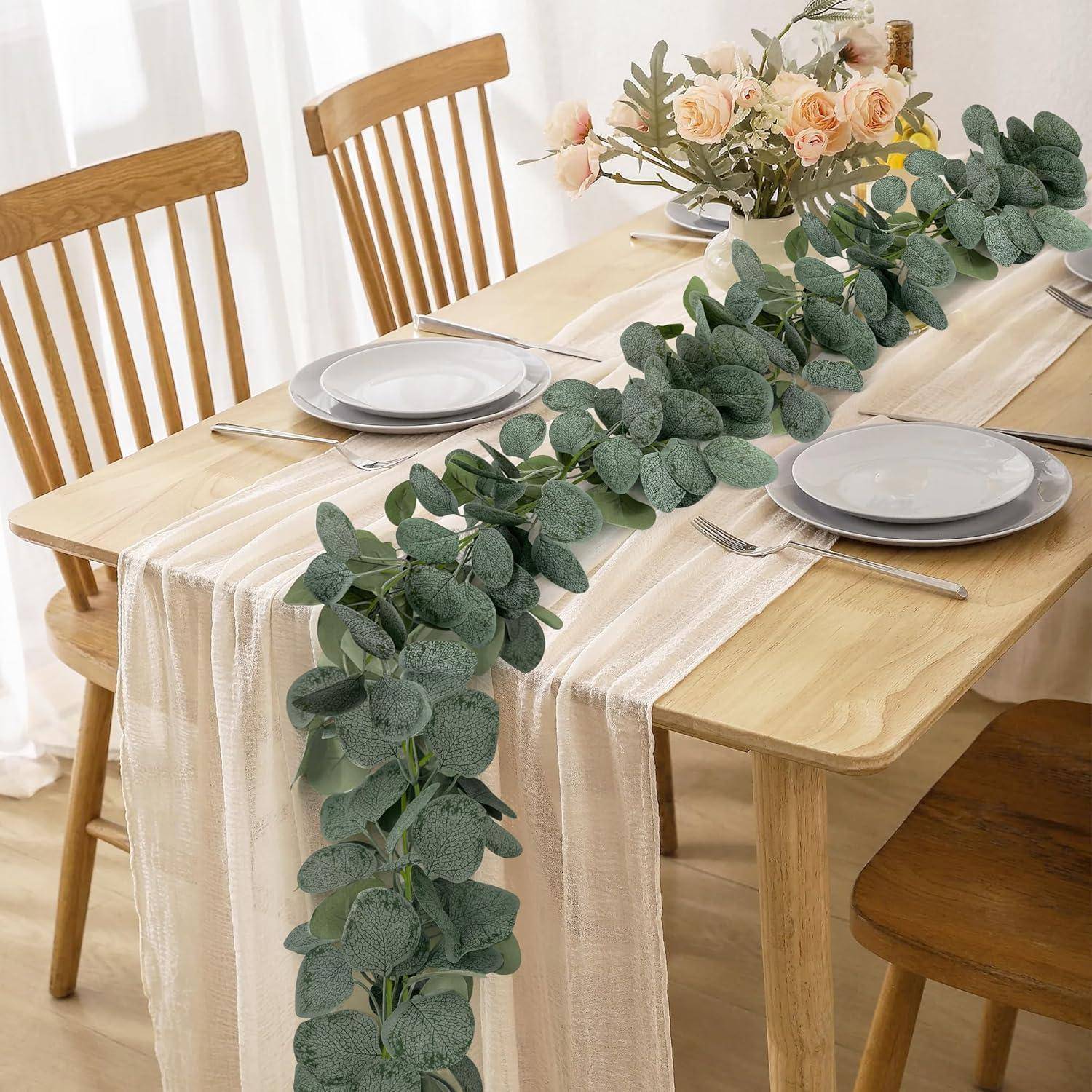 Ein Esstisch mit einem cremefarbenen Tischläufer, Tellern, Besteck und einer langen Blumenmitte aus Eukalyptus und Pfirsich, die eine stilvolle Mahlzeitsetting andeutet.