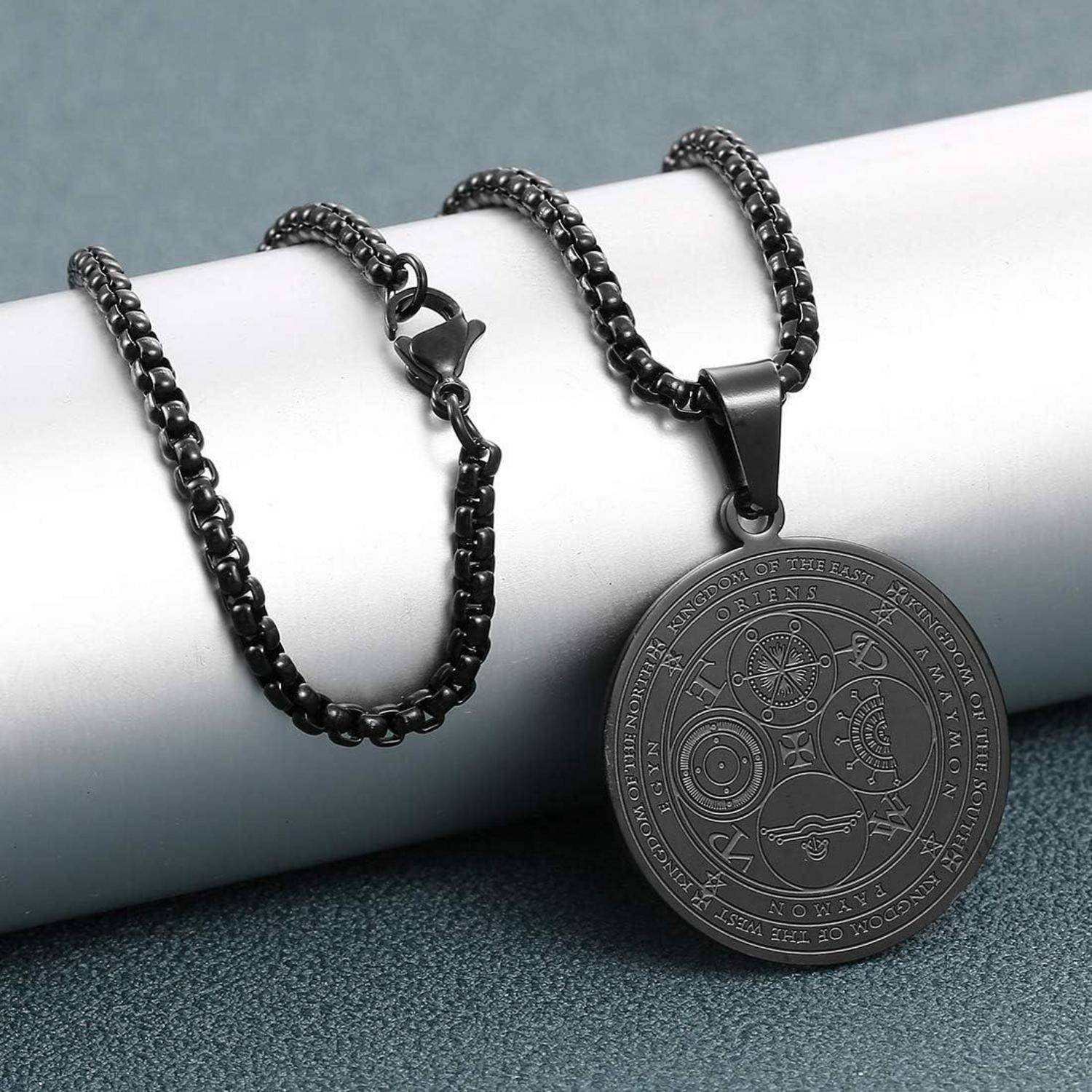 Schwarze Kettenkette mit rundem graviertem Anhänger, das auf einem hellen zylindrischen Display vor einem grauen Hintergrund ruht.
