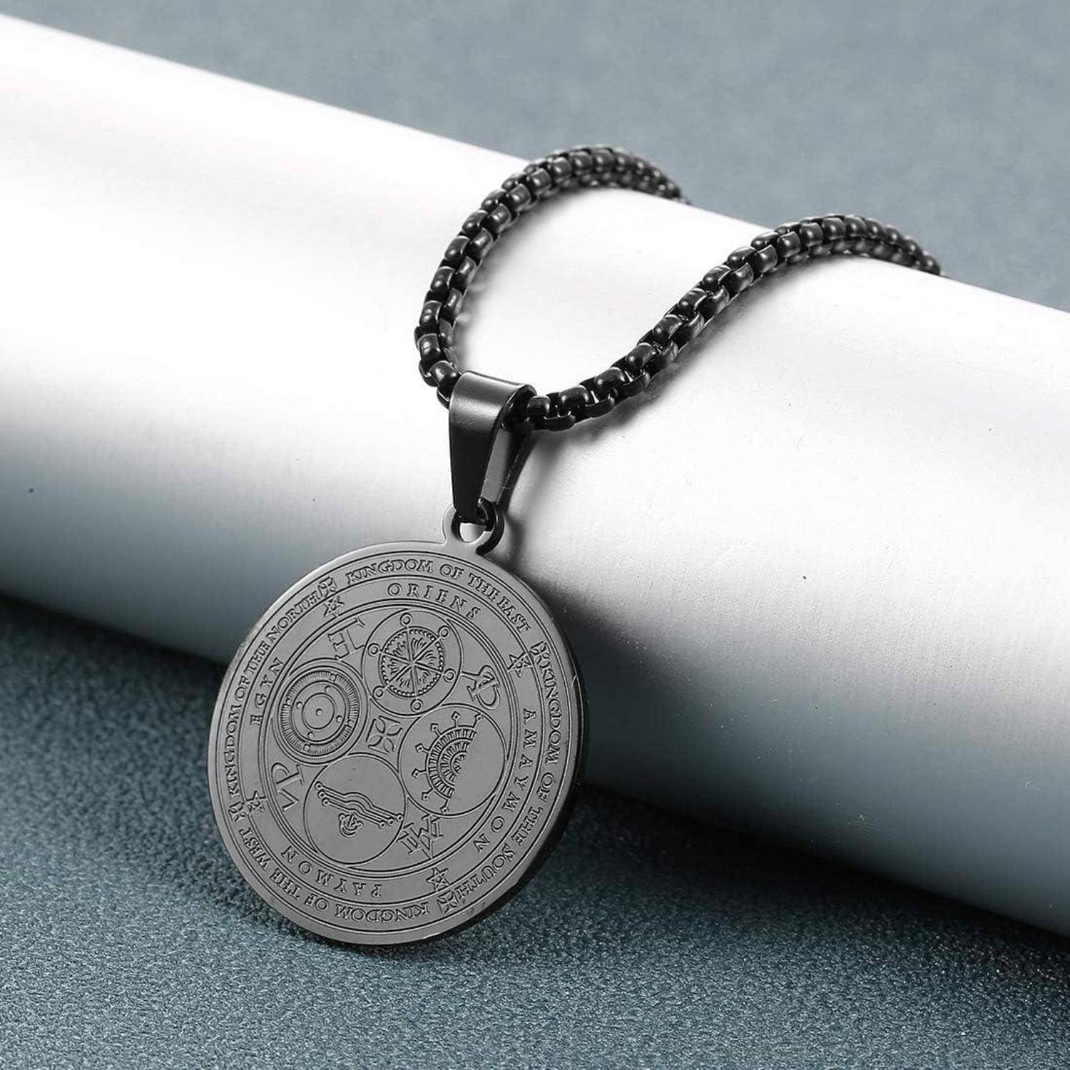 Schwarze Kettenkette mit einem runden gravierten Anhänger, der auf einem weißen Zylinder vor einem grauen Hintergrund ruht.