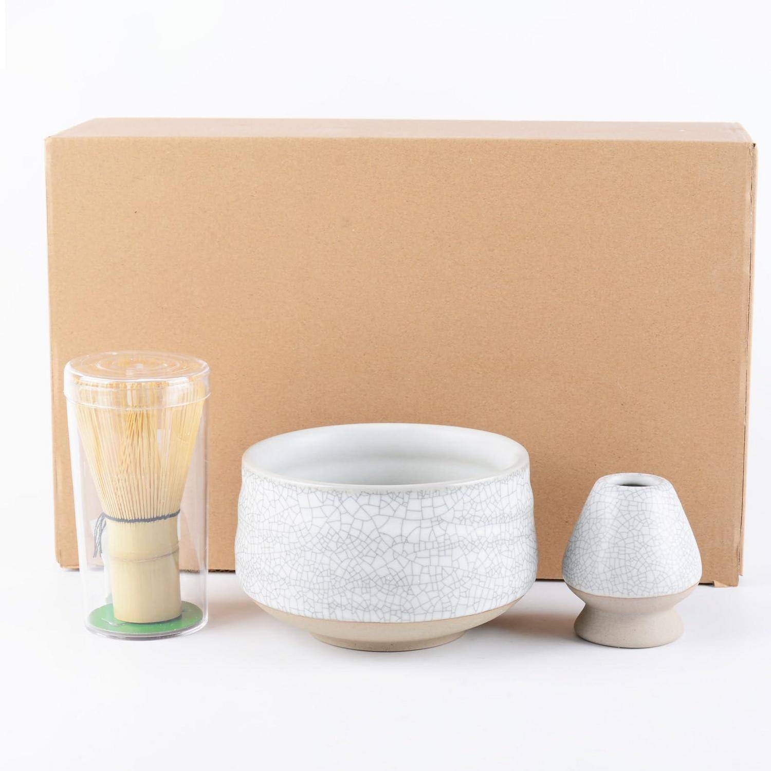 Weißes Keramik-Teeservice mit Bambusbesen, Schale und kleiner Tasse vor einem Karton auf weißem Hintergrund.