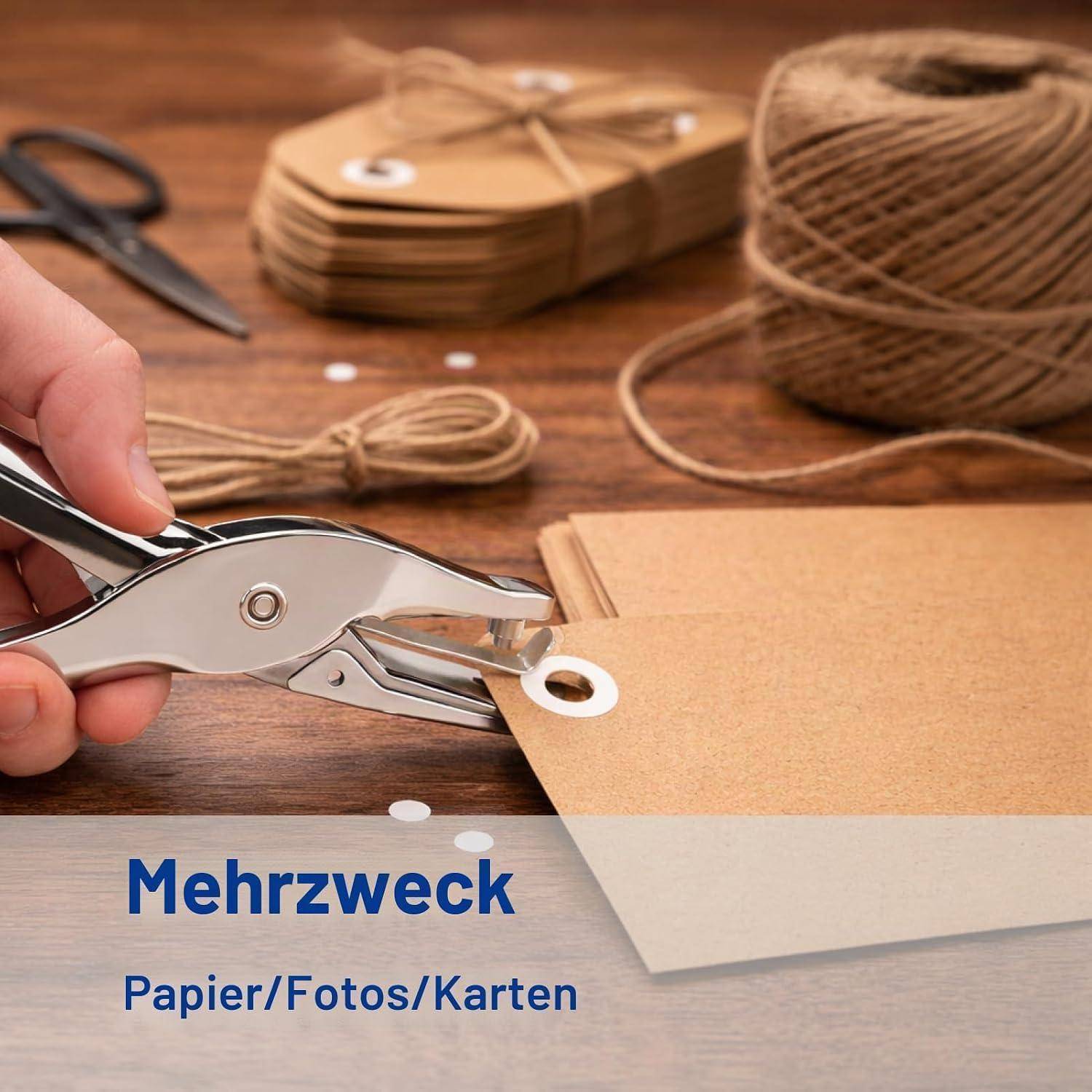 Hand benutzt ein Lochwerkzeug, um ein Loch in ein Kraftpapieretikett zu stanzen, mit Schnur, Schere und gestapelten Etiketten auf einem Holztisch; Text liest „Mehrzweck Papier/Fotos/Karten".