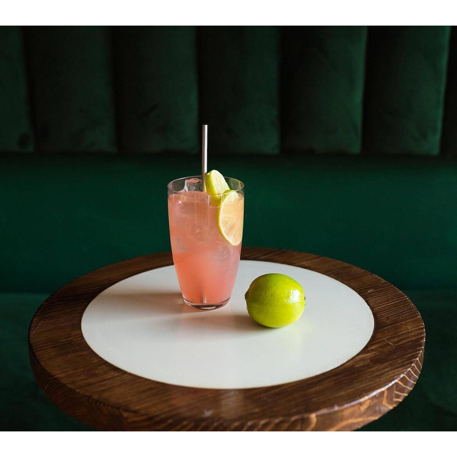 Pinkiger Cocktail in einem hohen Glas mit Strohhalm und Limettengarnierung, serviert auf einem runden Holztisch neben einer ganzen Limette.