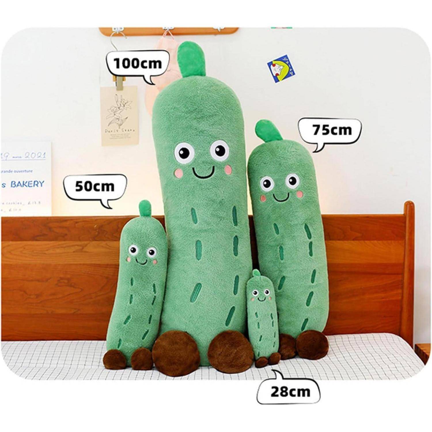 Vier lachende grüne Gurkenplüschtiere in verschiedenen Größen, beschriftet mit 100 cm, 75 cm, 50 cm und 28 cm, die auf einem Bett vor einem Holzkopfteil stehen.
