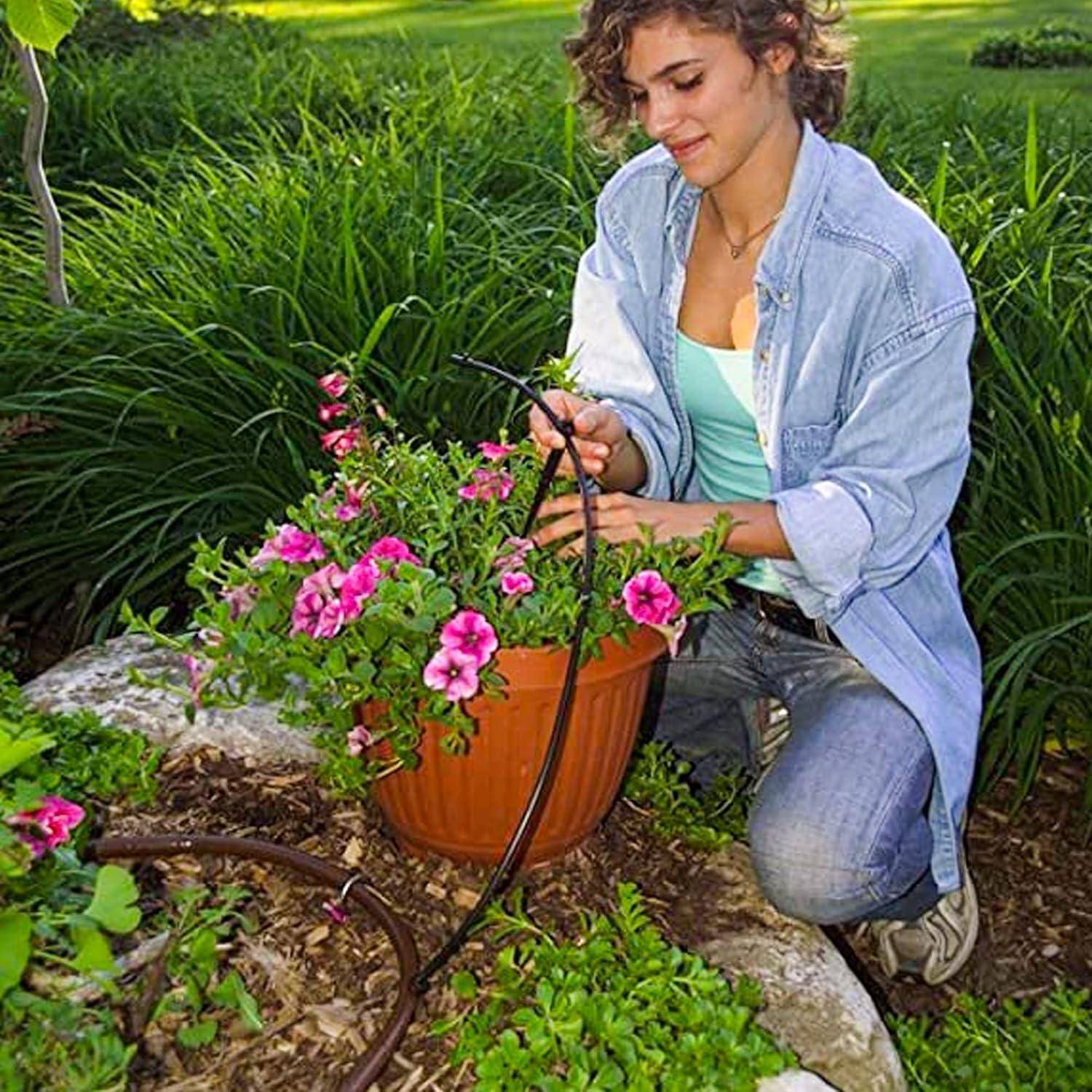 Frau kniet in einem Garten und bewässert rosa Blumen in einem Terrakottatopf mit einem Gartenschlauch.