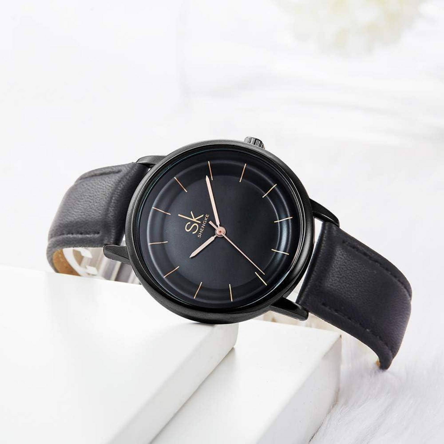 Schwarze analoge Armbanduhr mit Lederarmband, minimalen goldenen Stundenmarkern und 'SK'-Logo auf einem weißen Ausstellungsständer.