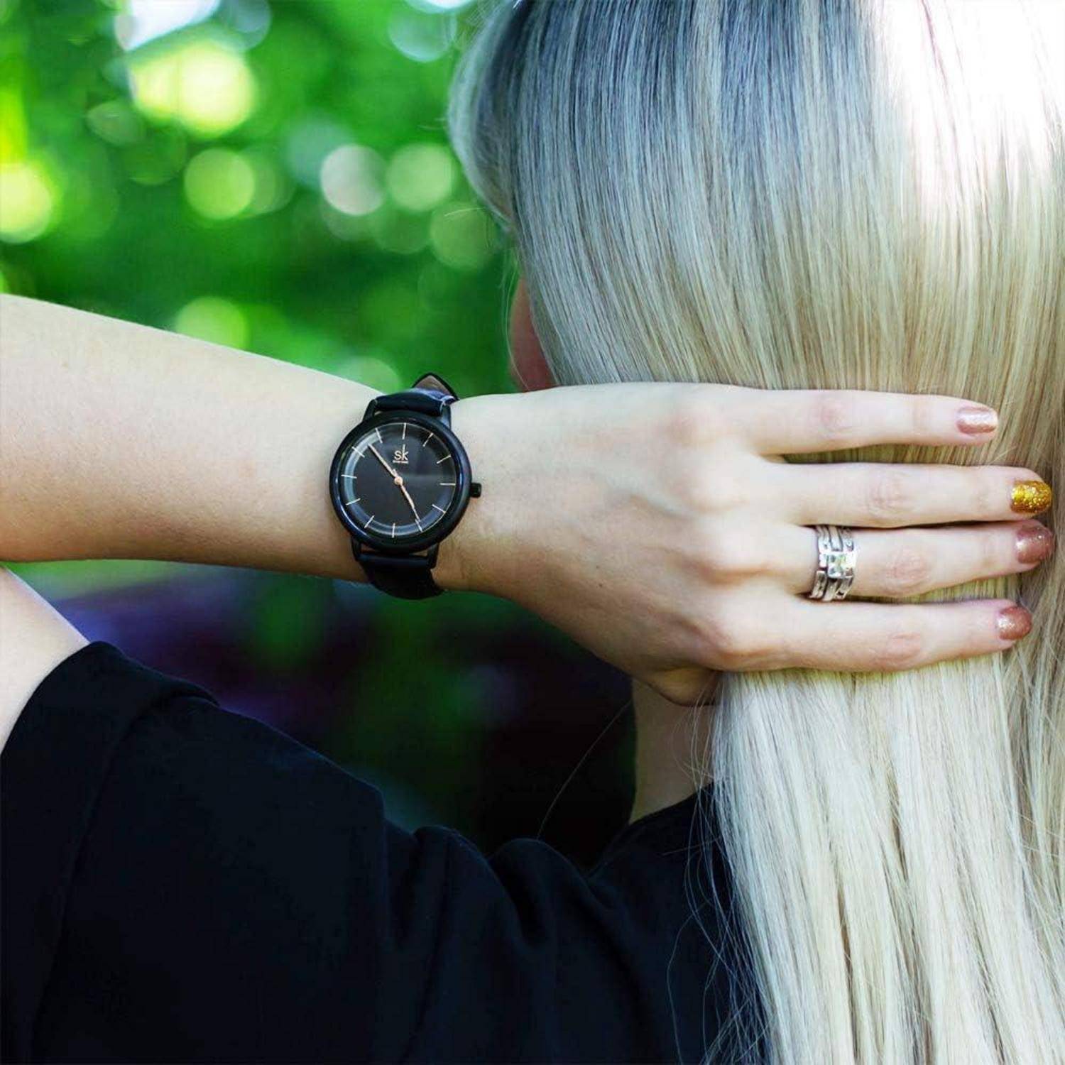 Nahaufnahme einer blonden Person von hinten, Hand berührt ihr Haar, trägt eine schwarze Armbanduhr und Ringe, vor einem grünem unscharfem Hintergrund.