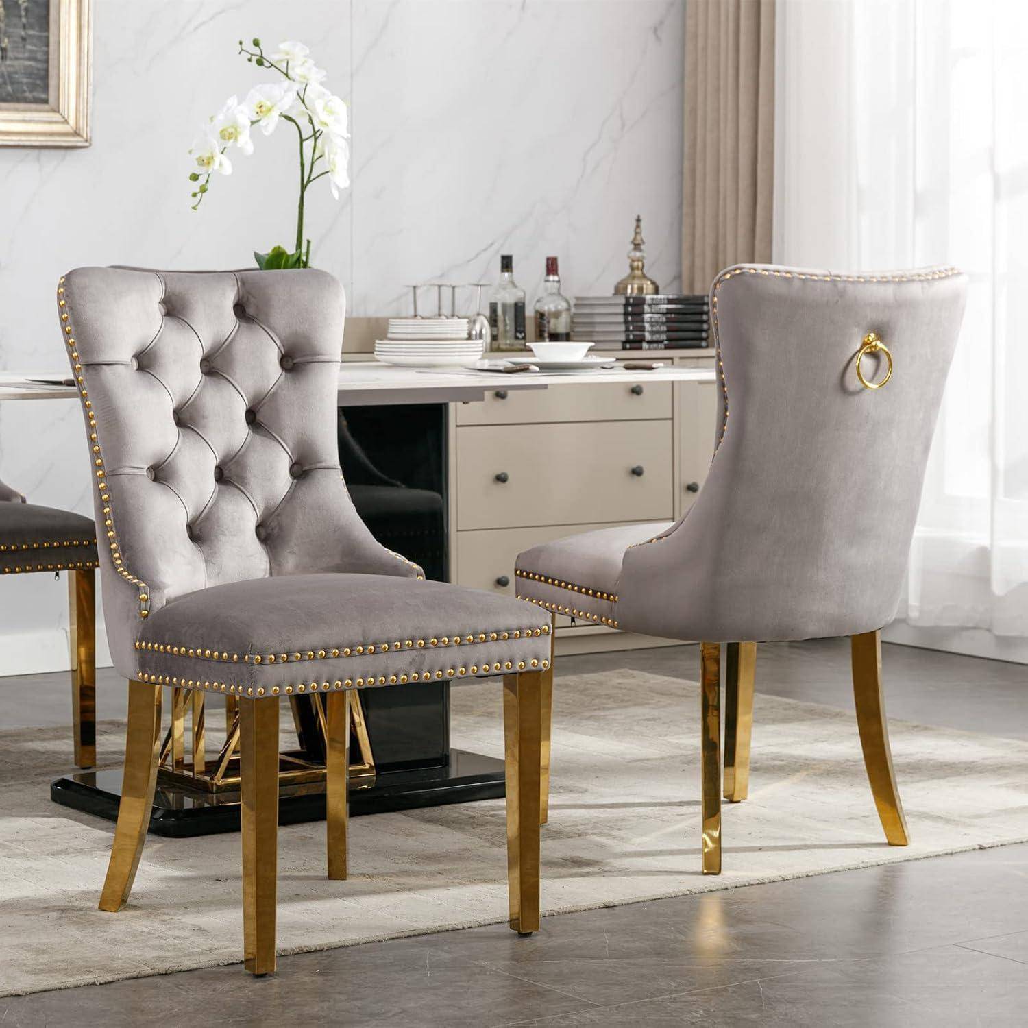 Zwei graue gepolsterte Esszimmerstühle mit Beinen aus Gold und Nagelkopfverzierung in einem modernen Zimmer neben einem weißen Schreibtisch und einer Orchidee.