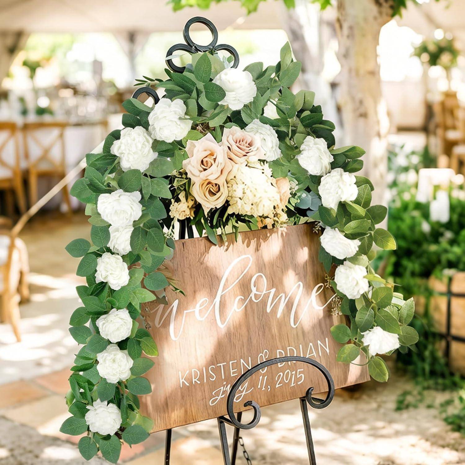 Willkommensschild mit Blumendekoration auf einer Staffelei, dekoriert mit weißen und hellrosa Blumen sowie Grünpflanzen; die Holzplakette trägt die Aufschrift „Willkommen" und „KRISTEN & BRIAN 11. Juli 2015"".