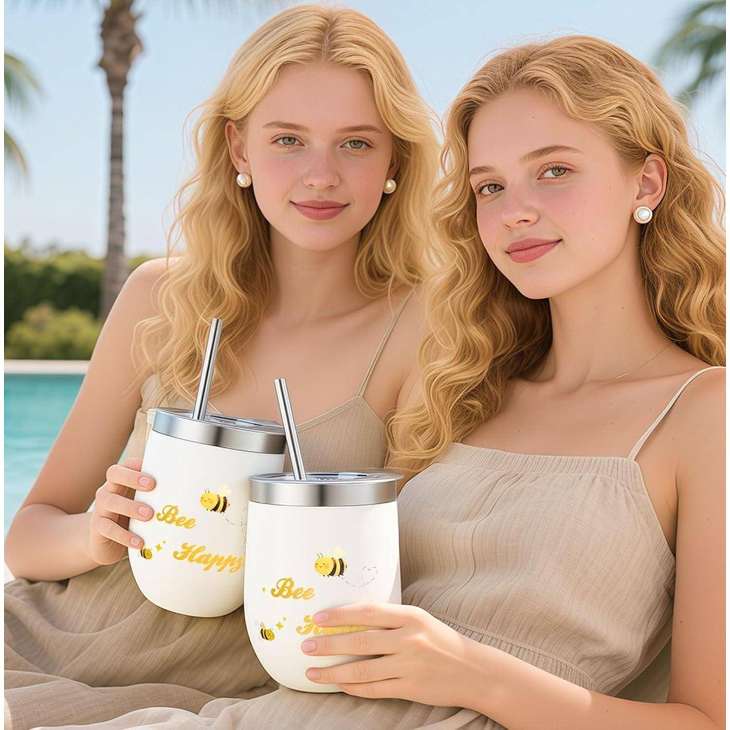 Zwei blonde Frauen in beigen Kleidern sitzen am Pool und halten übereinstimmende weiße Becher mit Bienen- und „Bee Happy
