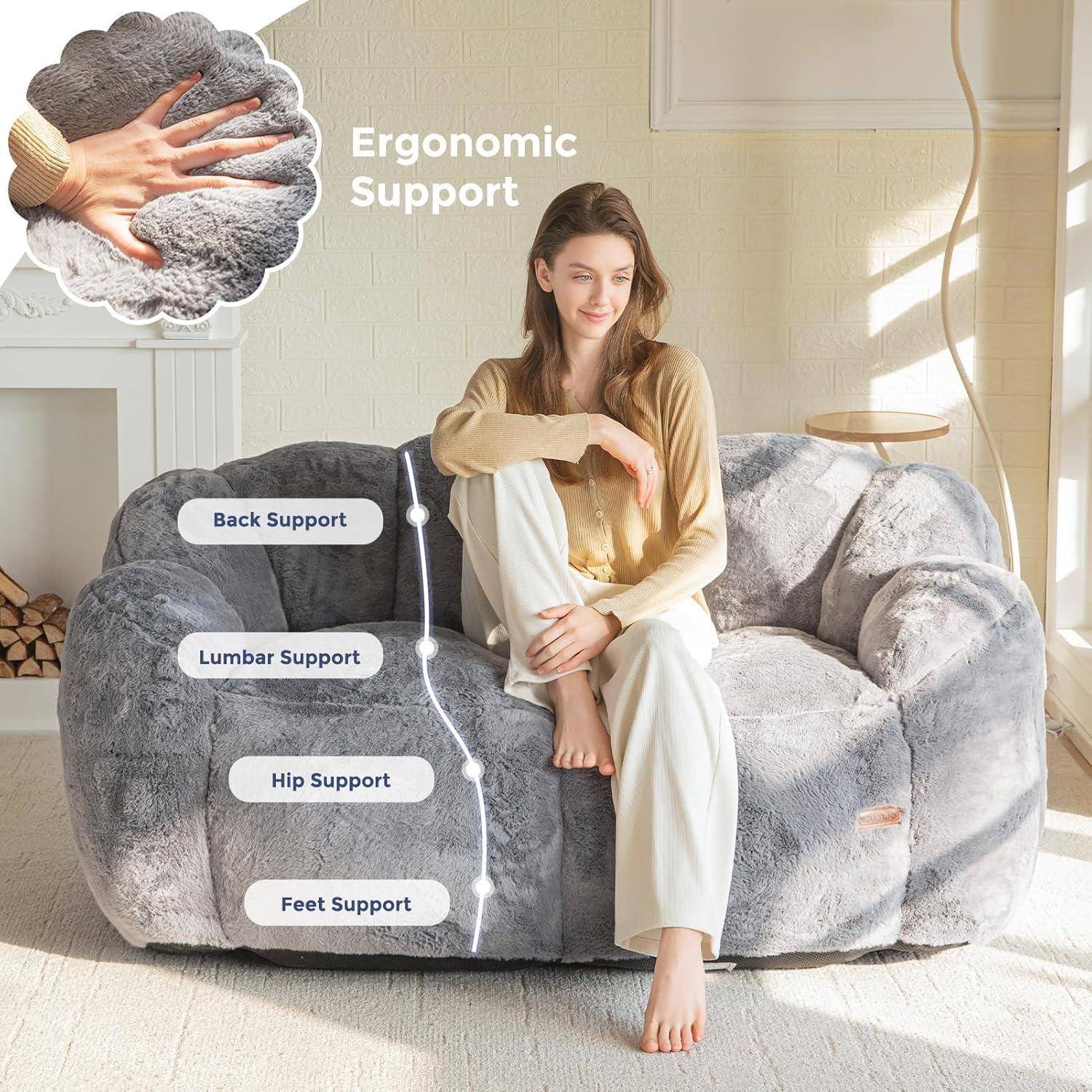 Frau sitzt auf einem luxuriösen grauen Sofa mit Beschriftungen für „Ergonomische Unterstützung", „Rückenstütze"", „Lendenwirbelstütze"", „Hüftstütze"" und „Fußstütze"".