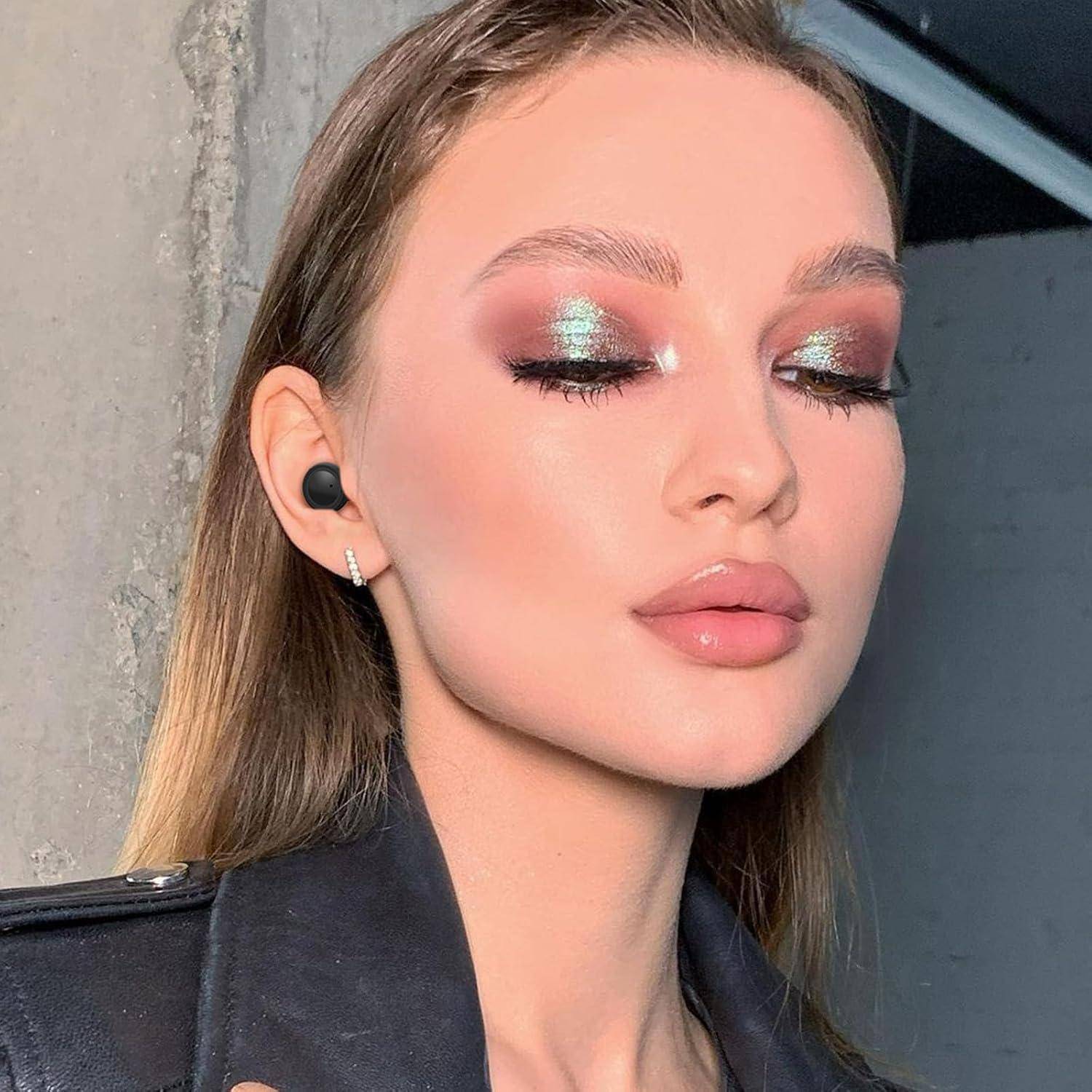 Nahaufnahme-Porträt einer Frau mit schimmerndem blaugrünem Lidschatten, glänzenden Lippen und einem schwarzen Ohrstöpsel, beleuchtet durch helles Sonnenlicht.