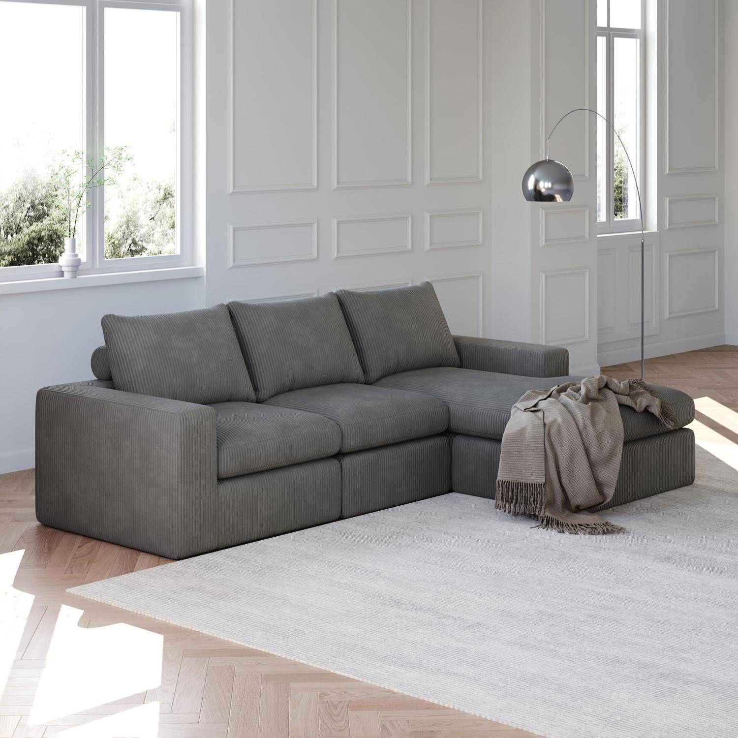 Modernes grau Eck-Sofa in einem hellen, minimalistischen Wohnzimmer mit einer Stehlampe, Wurfdecke, großen Fenstern und einem hellen Teppich.