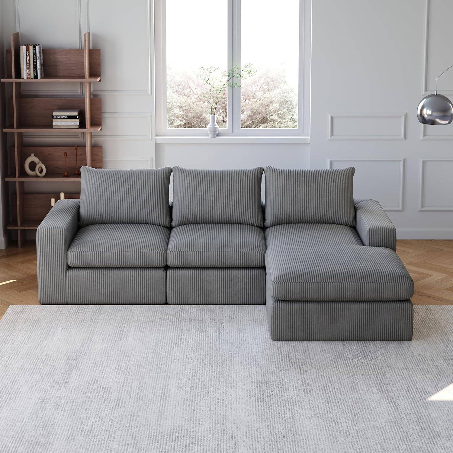 Graues Ecksofa mit Chaiselongue in einem hellen Wohnzimmer, an einer weißen Paneelwand und einem Fenster, mit Bücherregal und Stehlampe.